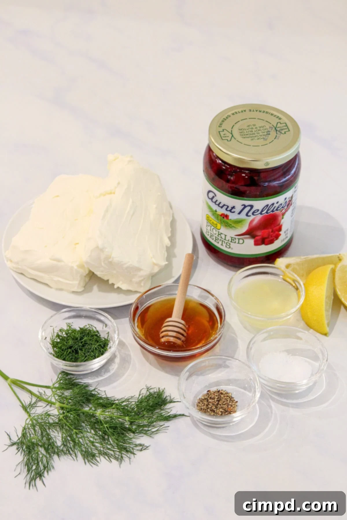 A collection of fresh ingredients for the beet cream cheese spread, including cream cheese, a jar of Aunt Nellie's diced pickled beets, honey, and fresh dill, arranged on a white marble counter.