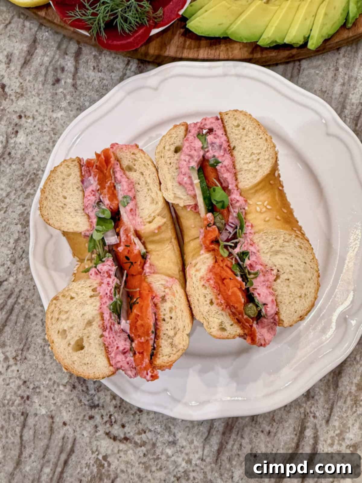 A delectable bagel sandwich, expertly cut in half, showcasing the layers of beet cream cheese spread, smoked salmon, and fresh vegetables on a white plate.