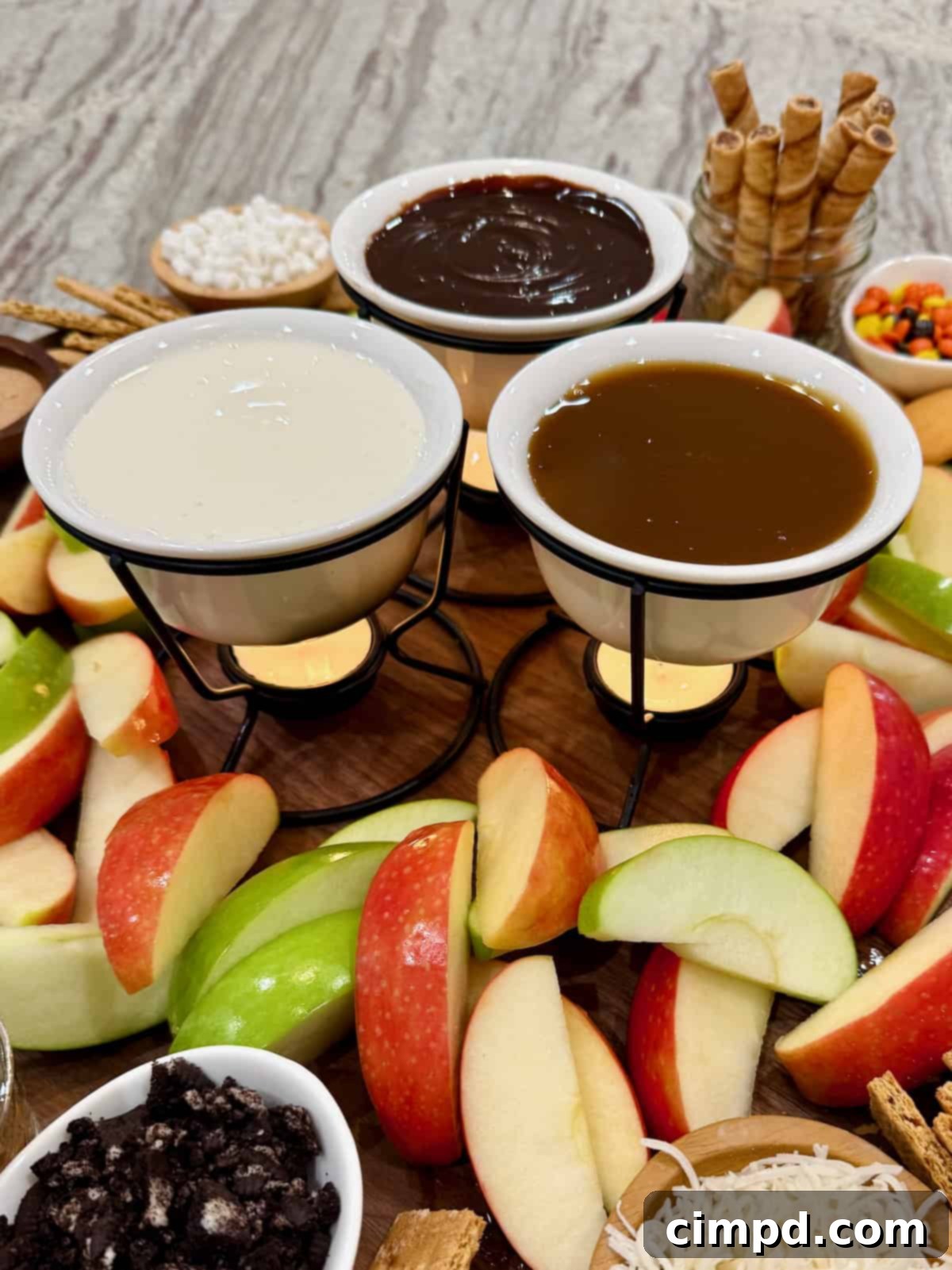 Fondue pots with hot fudge, marshmallow creme, and caramel sauce on a food board surrounded by apple slices.