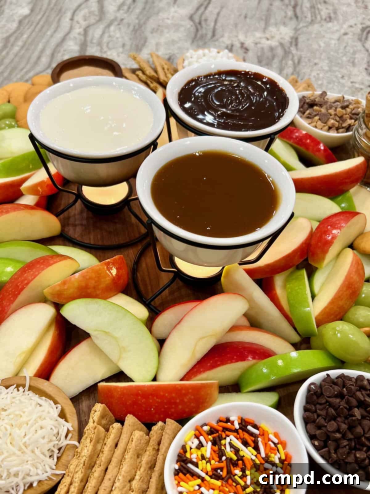 Fondue pots filled with hot fudge, marshmallow creme and caramel sauce on a food board surrounded by apple slices.