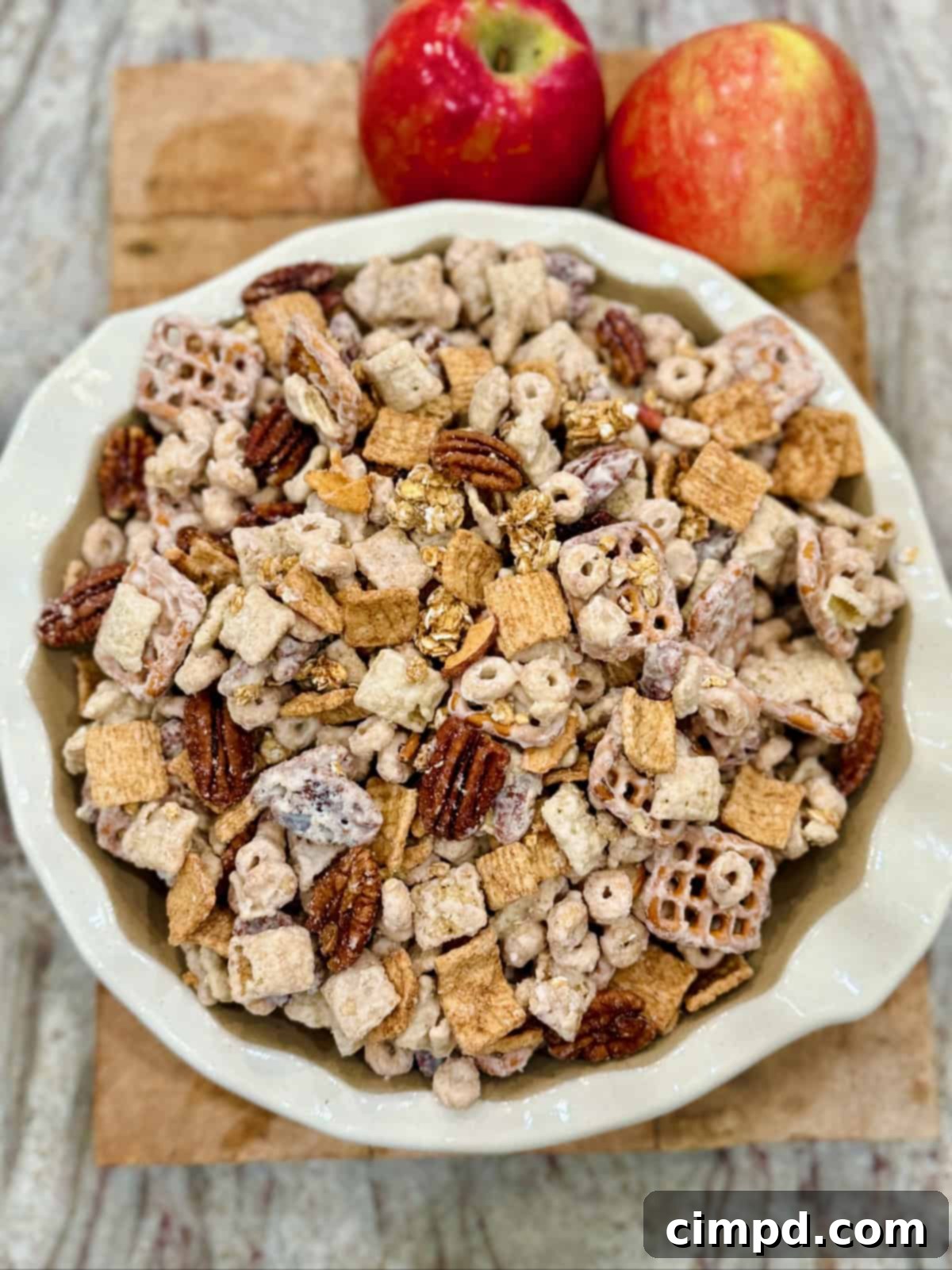 A beautifully arranged white scalloped bowl brimming with Apple Pie Snack Mix, set on a rustic wooden cutting board alongside two fresh, vibrant apples, evoking a sense of autumn and homemade goodness.