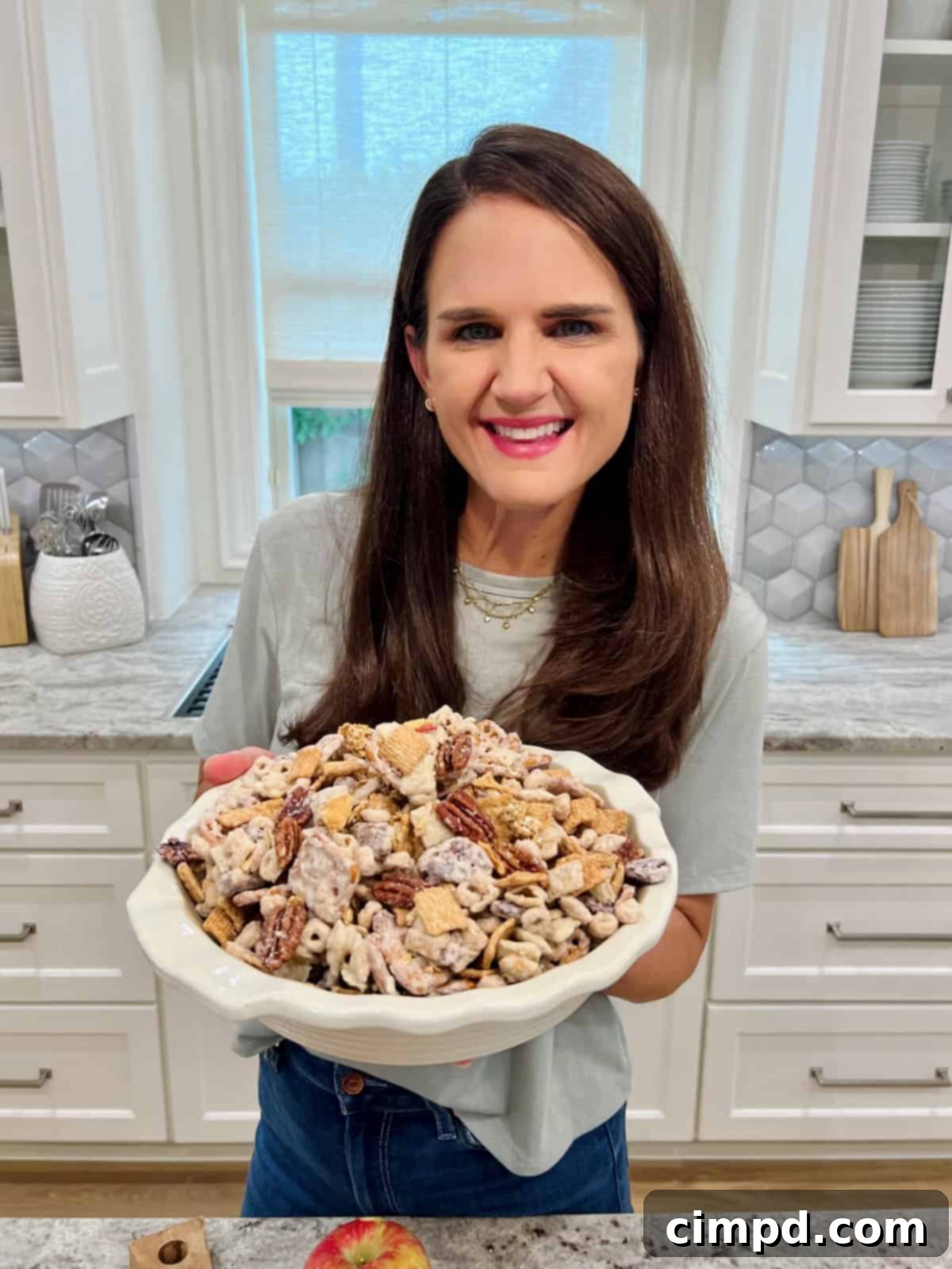 Maegan Brown, the BakerMama, warmly holding a white scalloped bowl overflowing with the freshly made Apple Pie Snack Mix, showcasing her pride and the snack's inviting appeal.