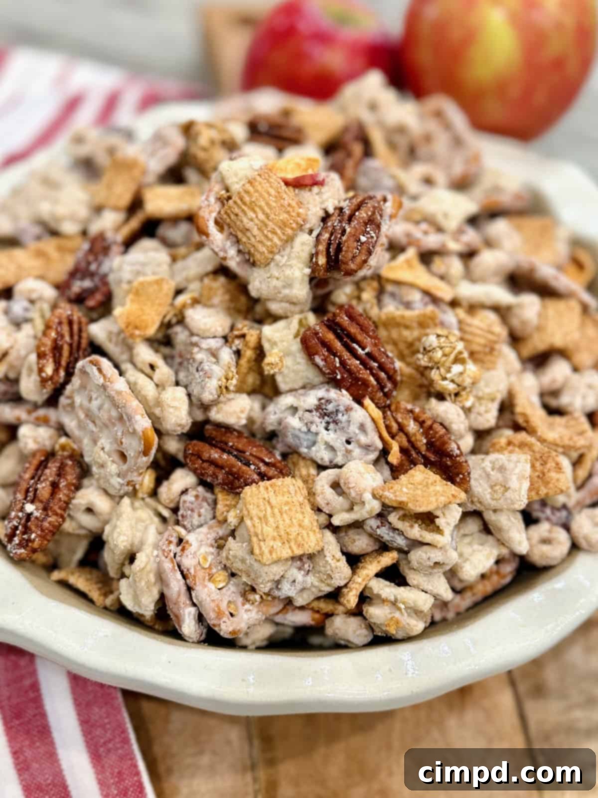 A pristine white scalloped bowl overflowing with the delicious Apple Pie Snack Mix, artfully placed on a wooden cutting board, with two fresh, green apples adding a touch of natural beauty and highlighting the snack's core flavor.