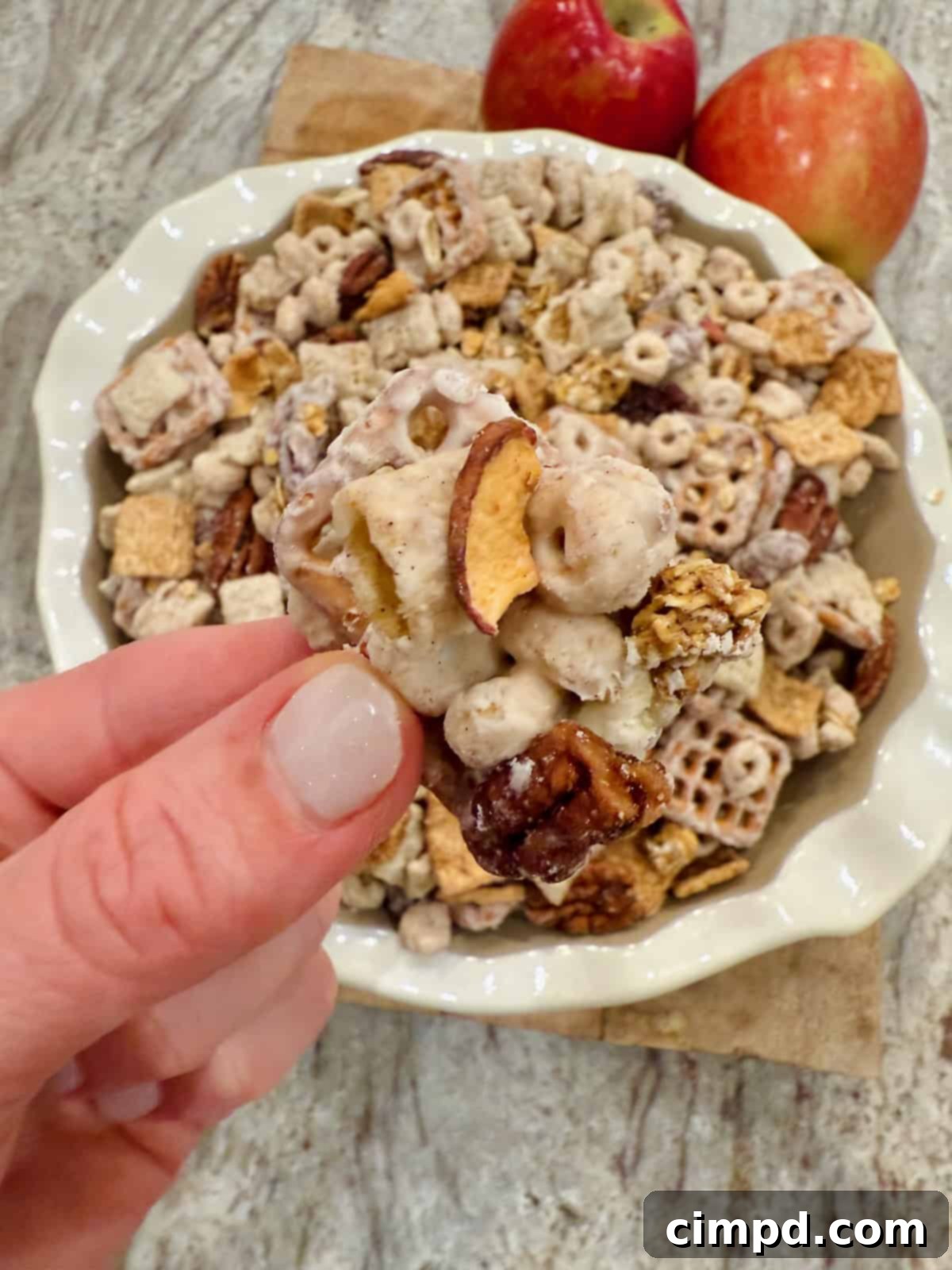 A close-up shot of a perfect cluster of Apple Pie Snack Mix held delicately between two fingers, highlighting the enticing candy coating and variety of ingredients.