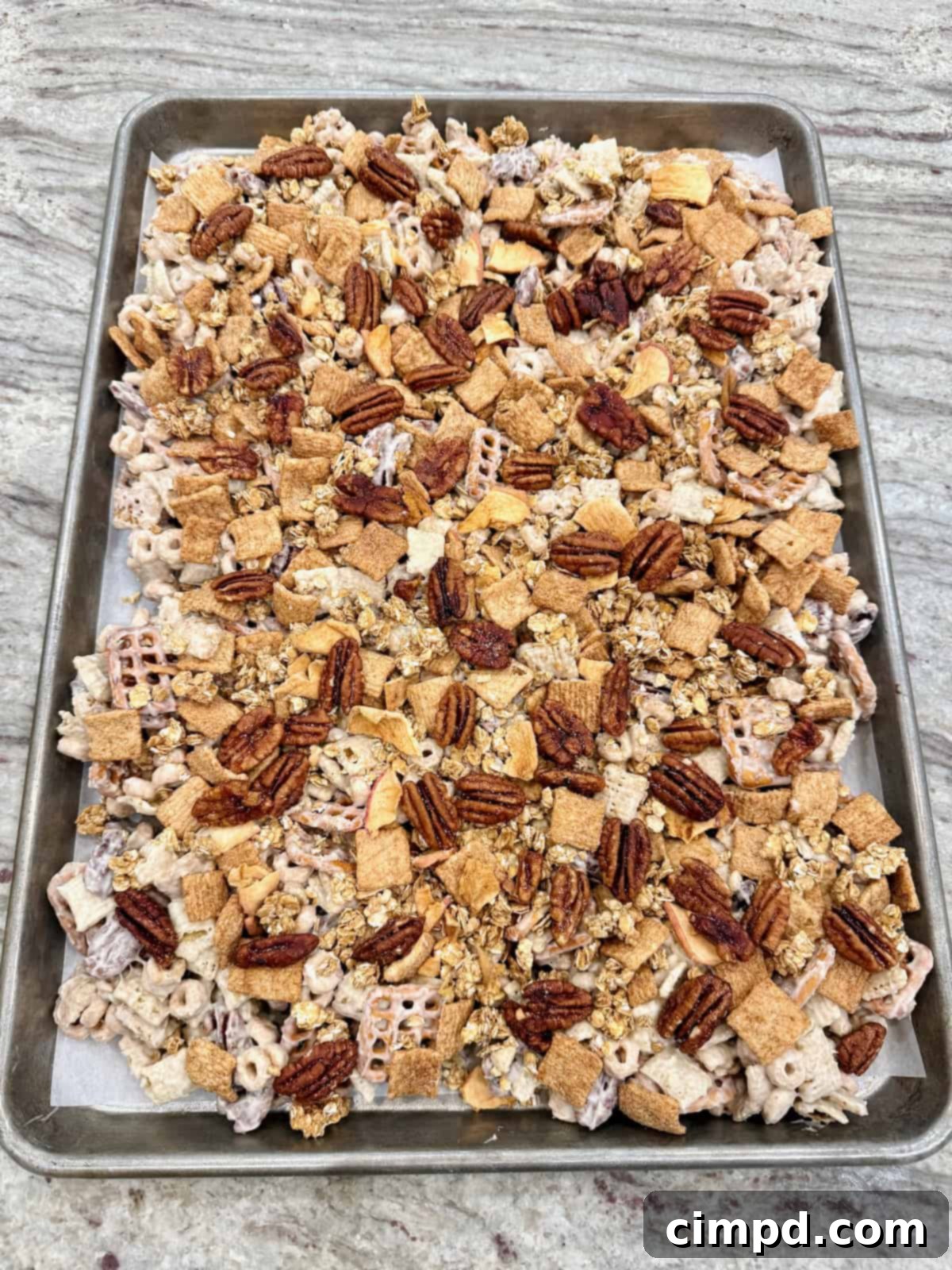 A large batch of freshly made Apple Pie Snack Mix spread out evenly on a parchment-lined baking sheet, glistening with its candy coating and ready to be broken into clusters.