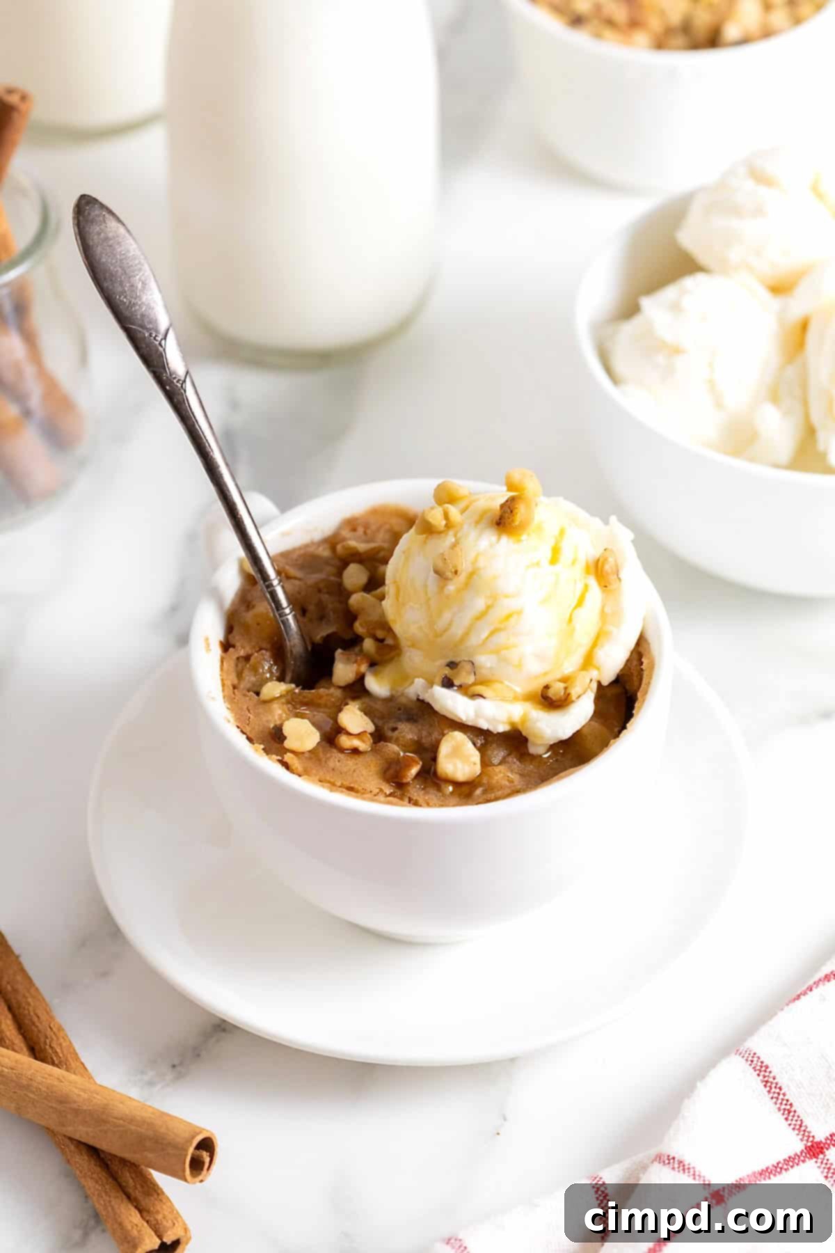 Spiced Apple Mug Delight 5 A freshly baked apple mug cake, adorned with a melting scoop of vanilla ice cream, a spoon resting enticingly within. It's served on a white plate, ready to be enjoyed.