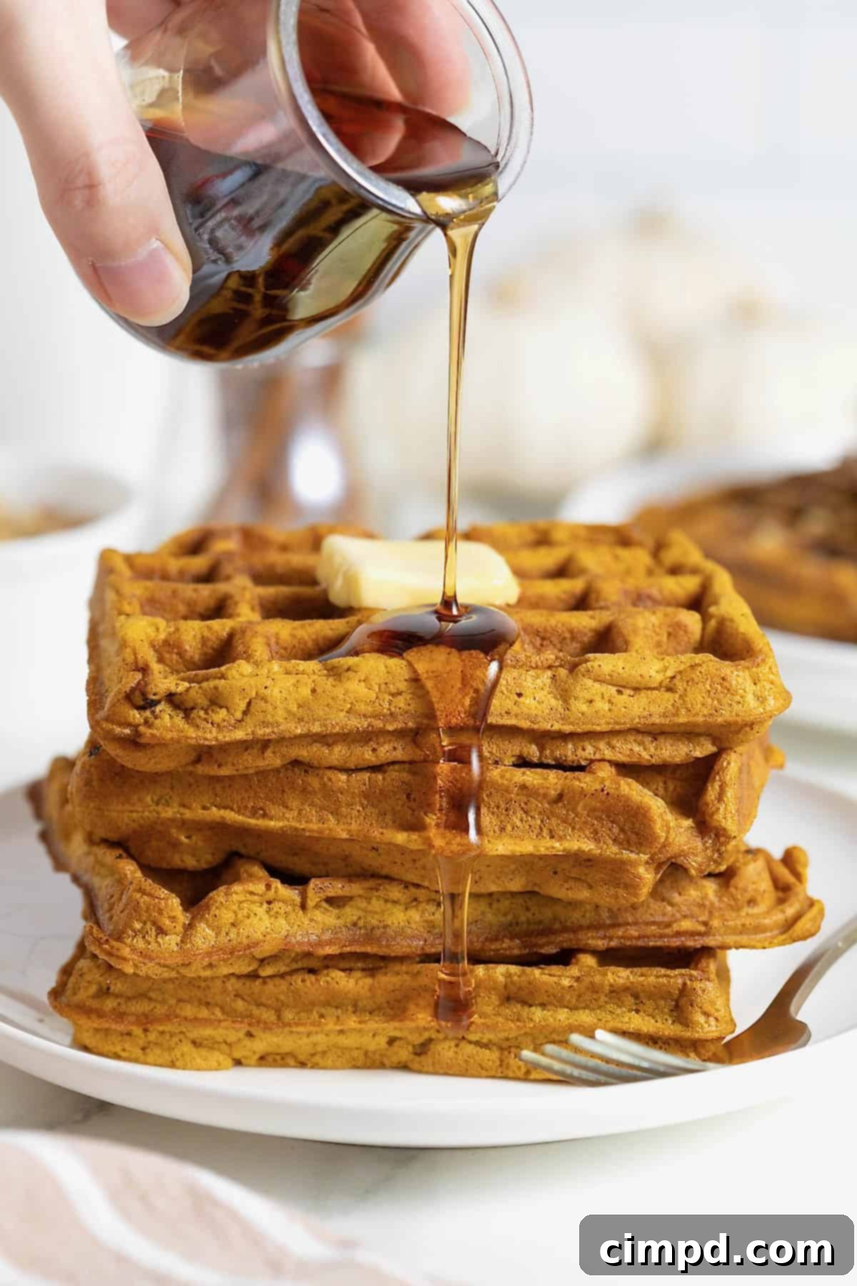 Spiced Pumpkin Waffles 4 Golden maple syrup being generously poured over a delectable stack of five fluffy pancakes on a white plate, ready to be enjoyed.