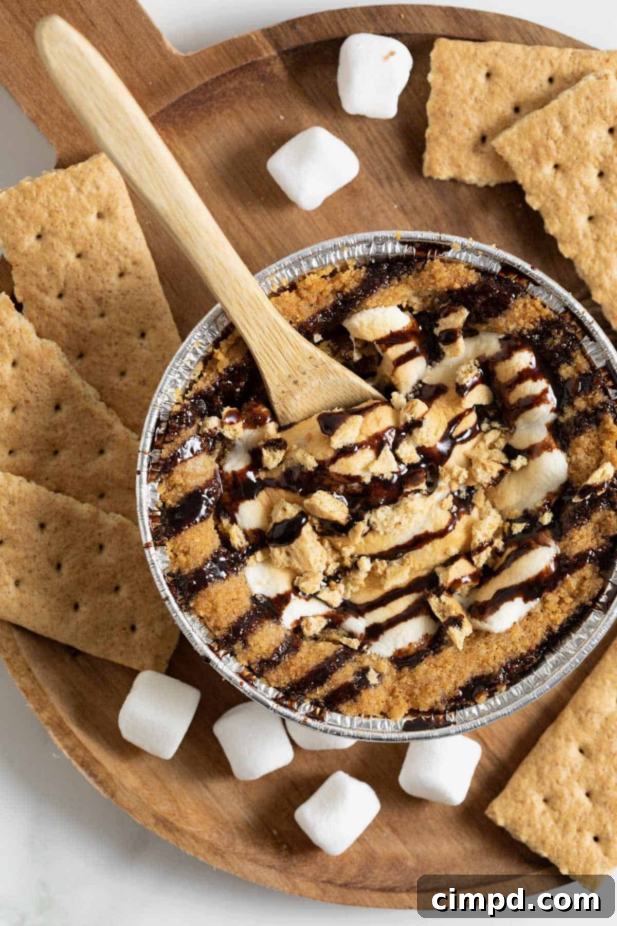 A perfectly baked mini s'mores cookie pie, golden brown with toasted marshmallows, served on a rustic wooden cutting board. Mini marshmallows and broken graham cracker pieces are scattered around, inviting a taste.