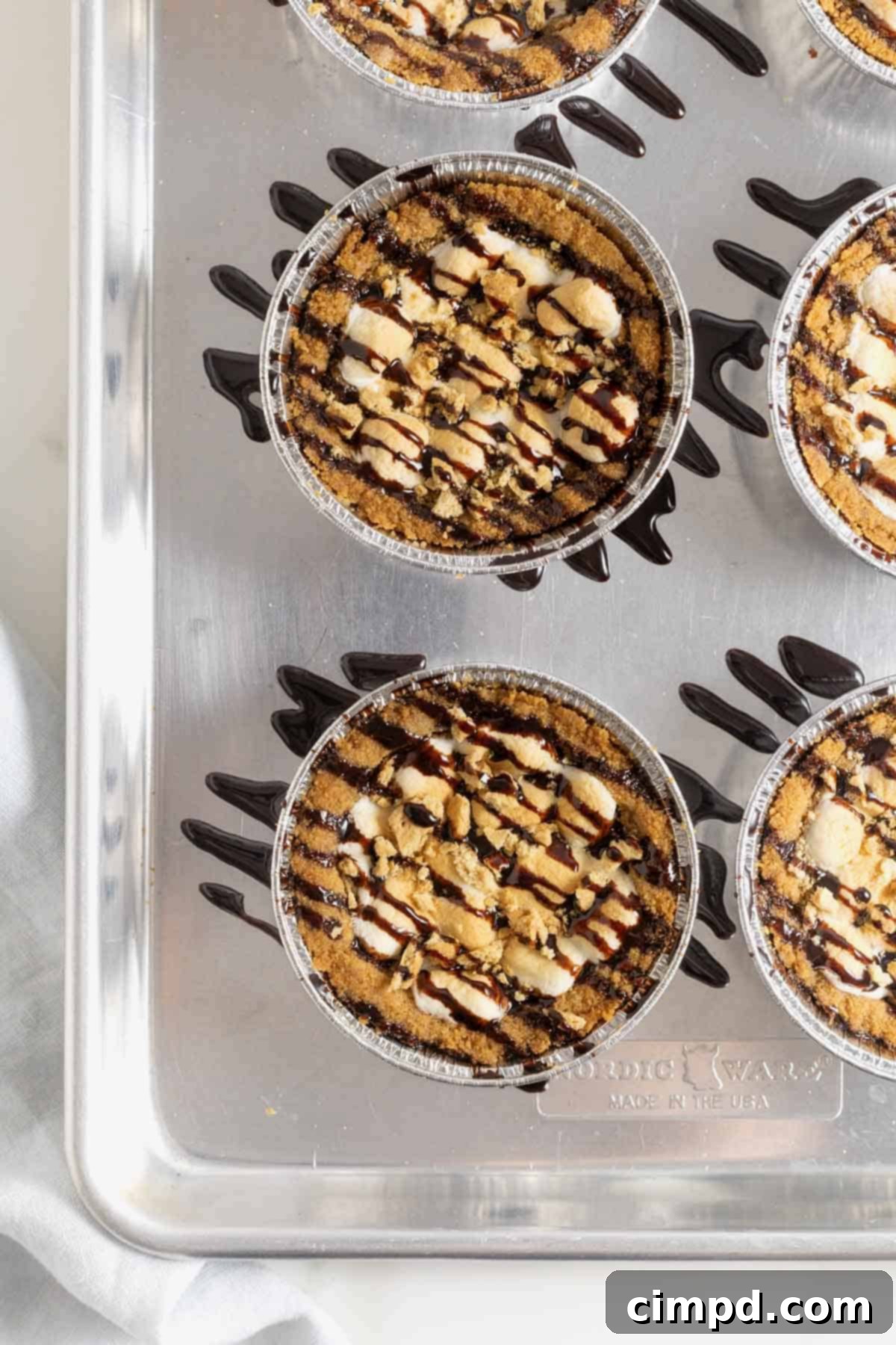 Five mini s'mores cookie pies, drizzled generously with chocolate sauce, arranged on an aluminum baking sheet, ready to be served or stored.