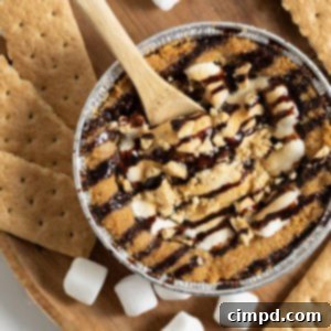 A mini s'mores cookie pie on a wooden cutting board surrounded by mini marshmallows and graham cracker pieces, representing the final delicious product of the recipe.