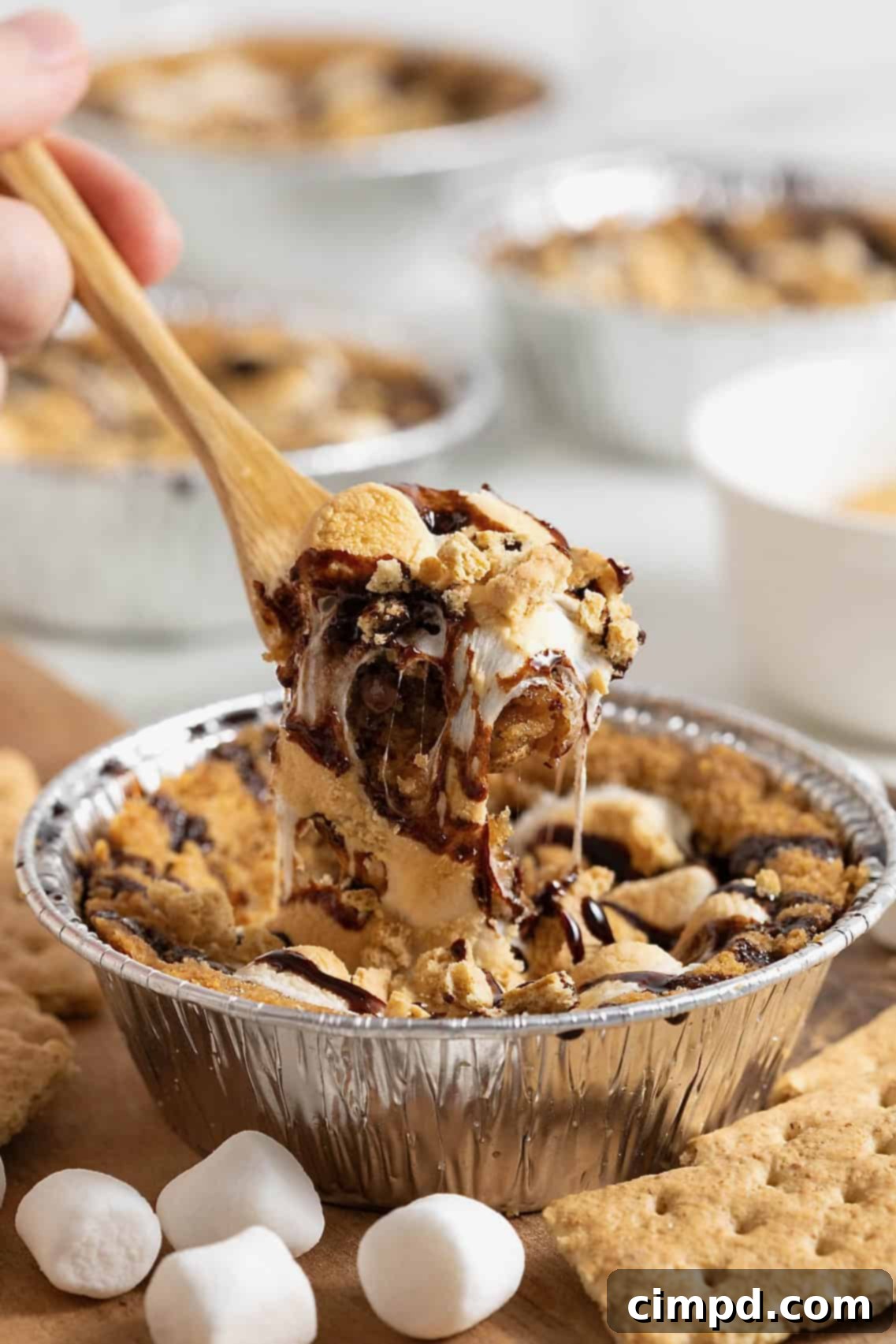 A wooden spoon carefully lifts a serving of a mini s'mores cookie pie, showcasing its golden crust, melted cookie dough, and perfectly gooey, toasted marshmallow topping, ready to be enjoyed.