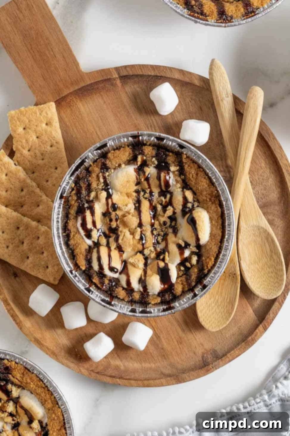 A single mini s'mores cookie pie, with its golden crust, melted chocolate chip cookie, and perfectly toasted marshmallows, rests on a small round wooden board, surrounded by mini marshmallows, graham cracker pieces, and two decorative wooden spoons.
