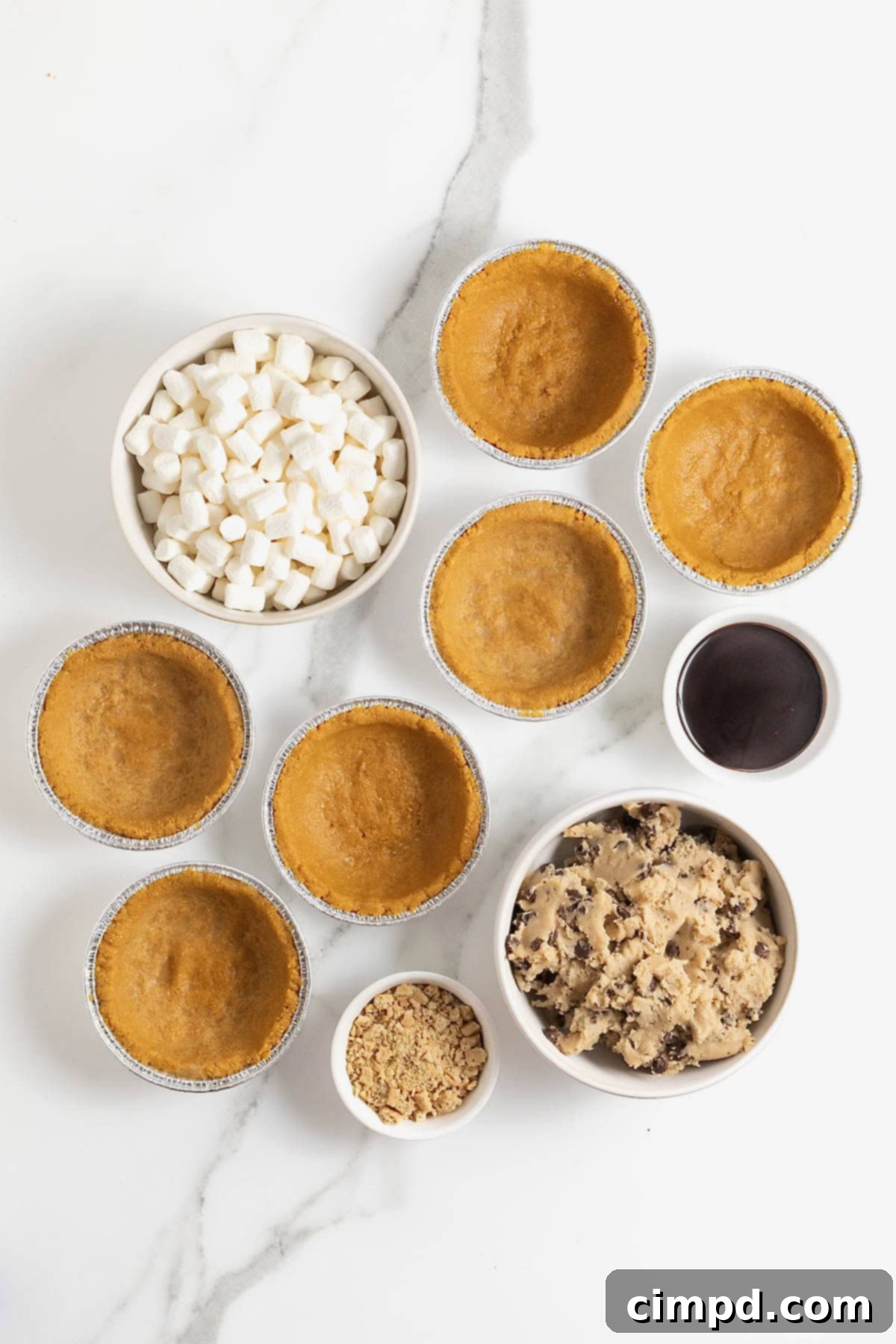 A selection of ingredients laid out on a white marble counter for making mini s'mores cookie pies, including mini graham cracker crusts, chocolate chip cookie dough, a bag of mini marshmallows, and bottles of chocolate syrup.