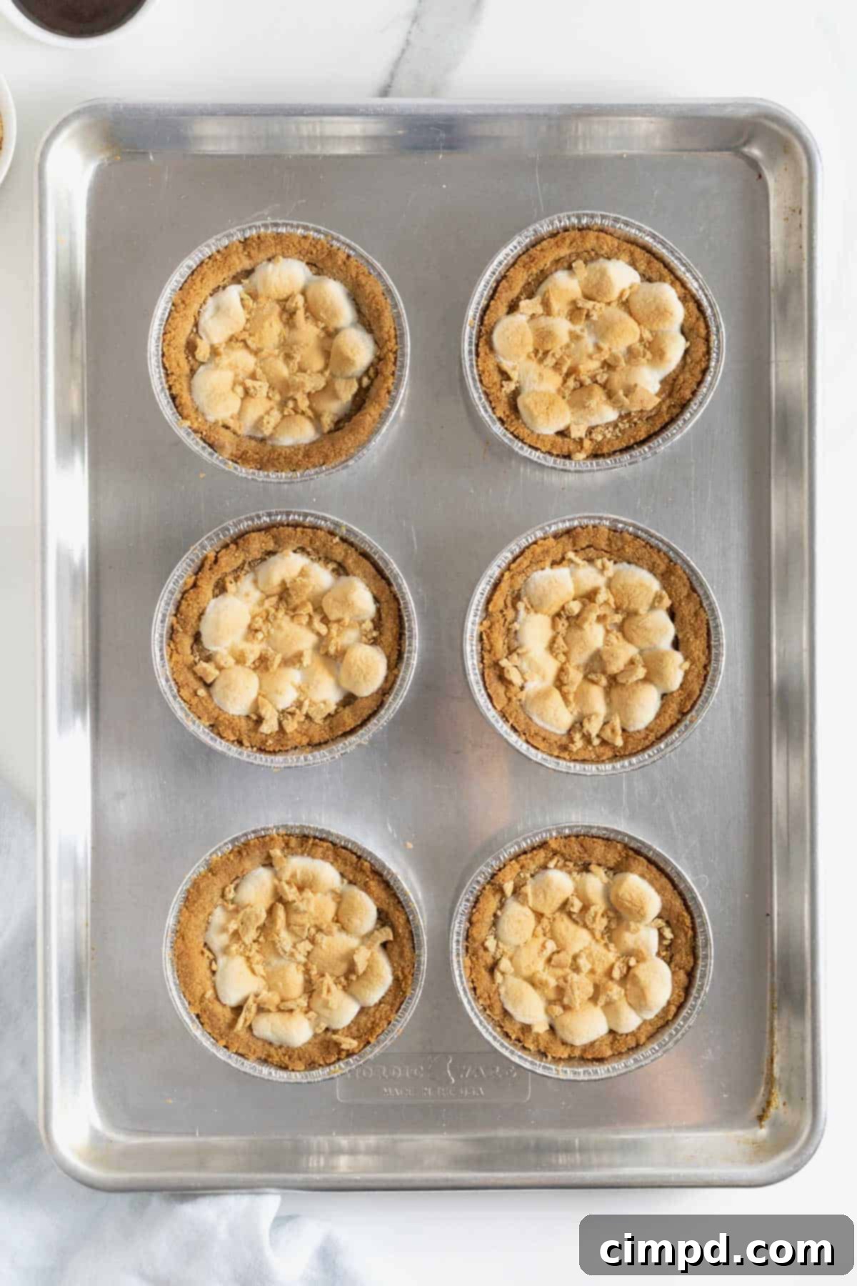 Six freshly baked s'mores cookie pies, still on the aluminum baking sheet, beautifully topped with a generous sprinkle of graham cracker crumbs, awaiting their final chocolate drizzle.