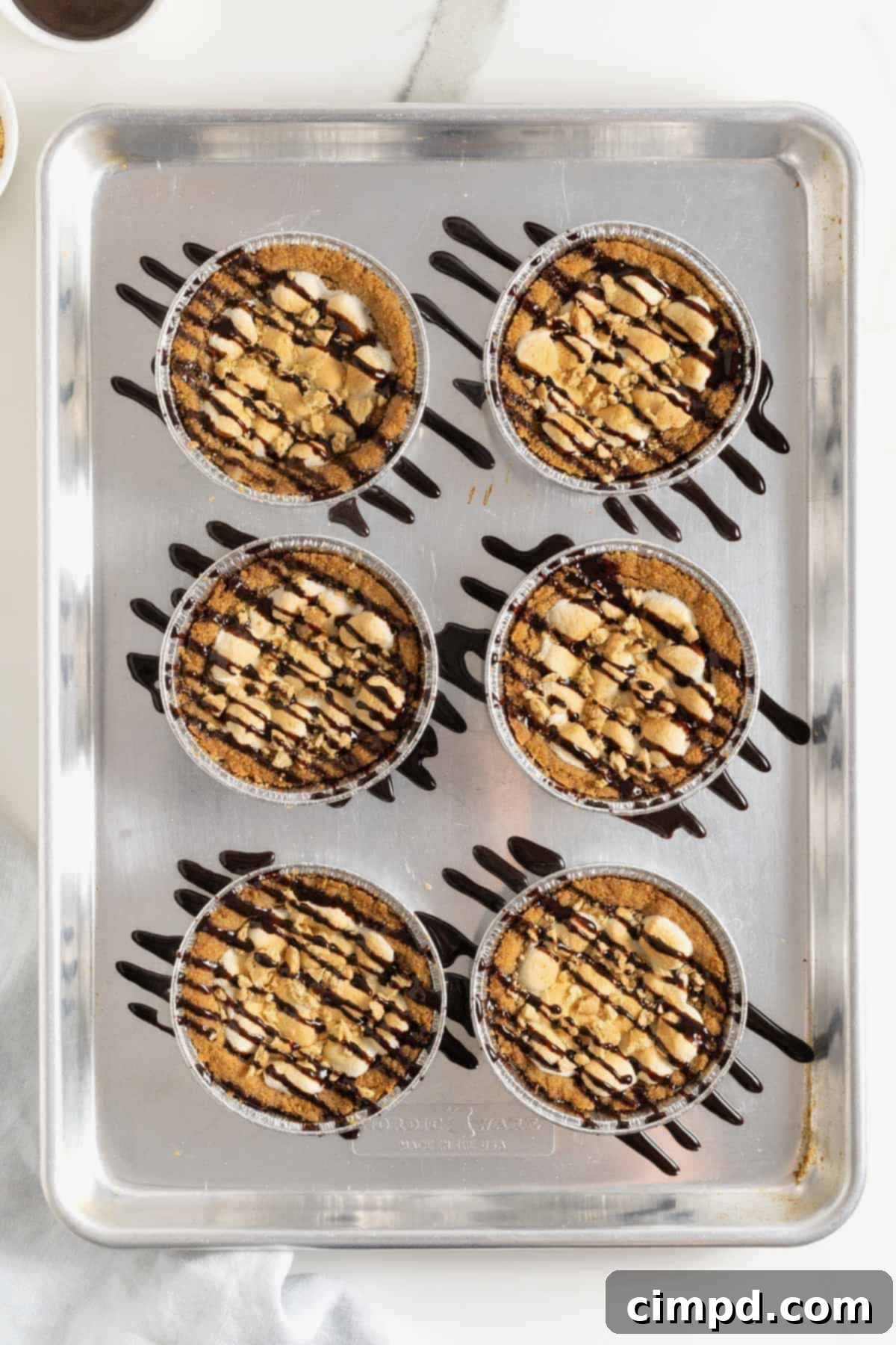 Six warm mini s'mores cookie pies, freshly drizzled with rich chocolate sauce and sprinkled with graham cracker crumbs, arranged on an aluminum baking sheet, ready for serving.