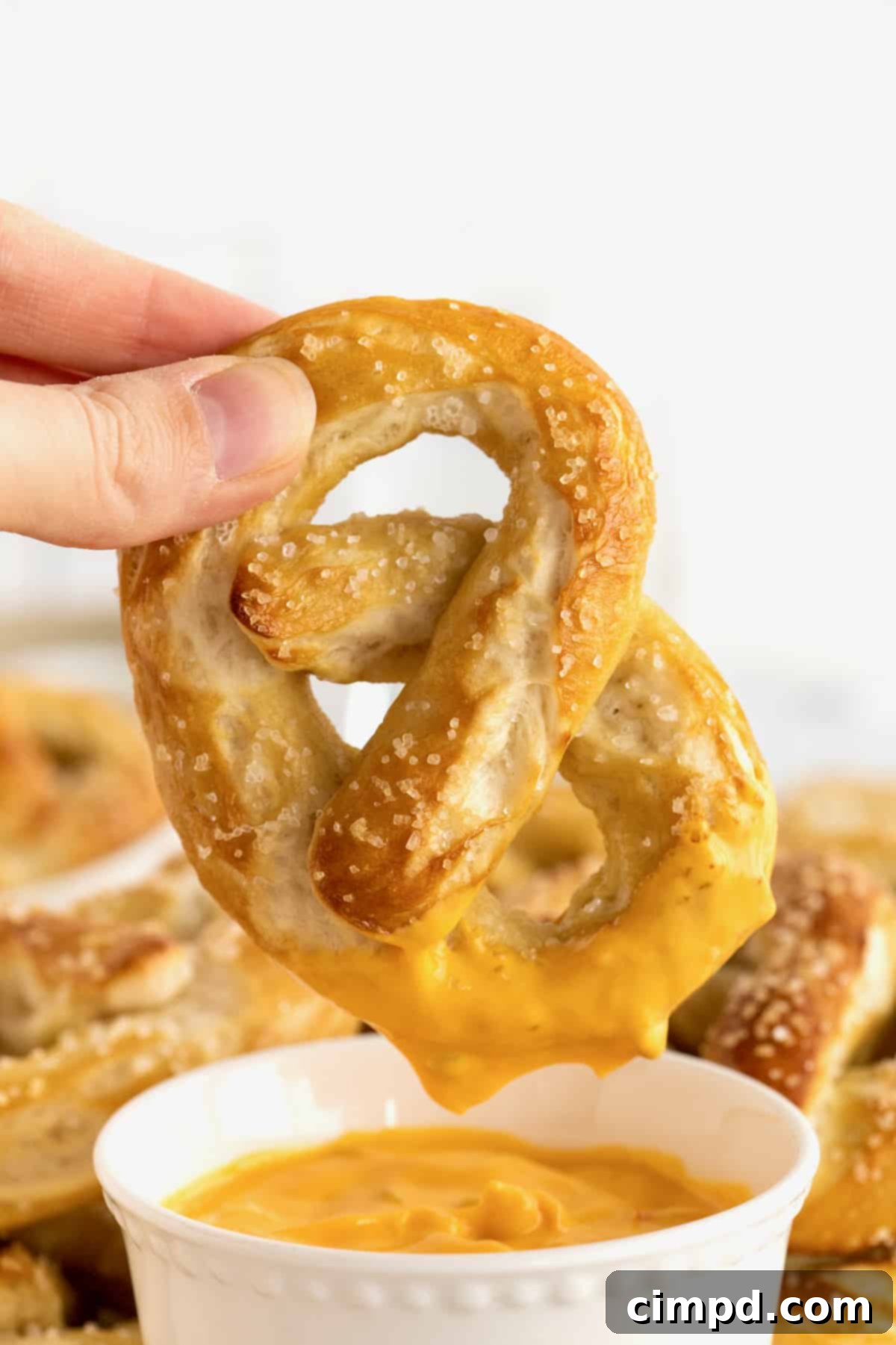 A hand holding a warm, soft pretzel just dipped into a rich, golden cheese sauce, with more pretzels and sauce blurred in the background.
