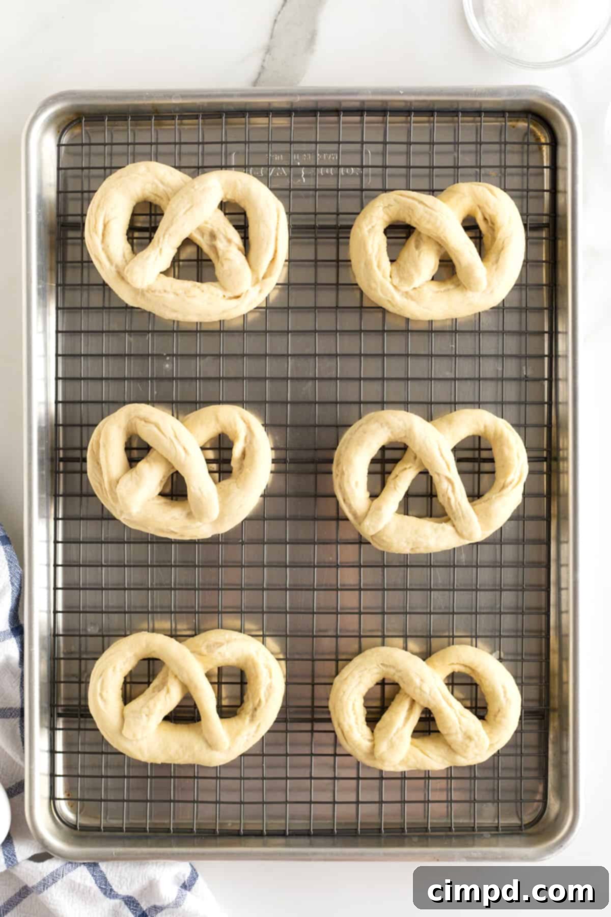 Six uncooked soft pretzels, perfectly shaped and ready for baking, resting on a wire rack over a baking sheet.