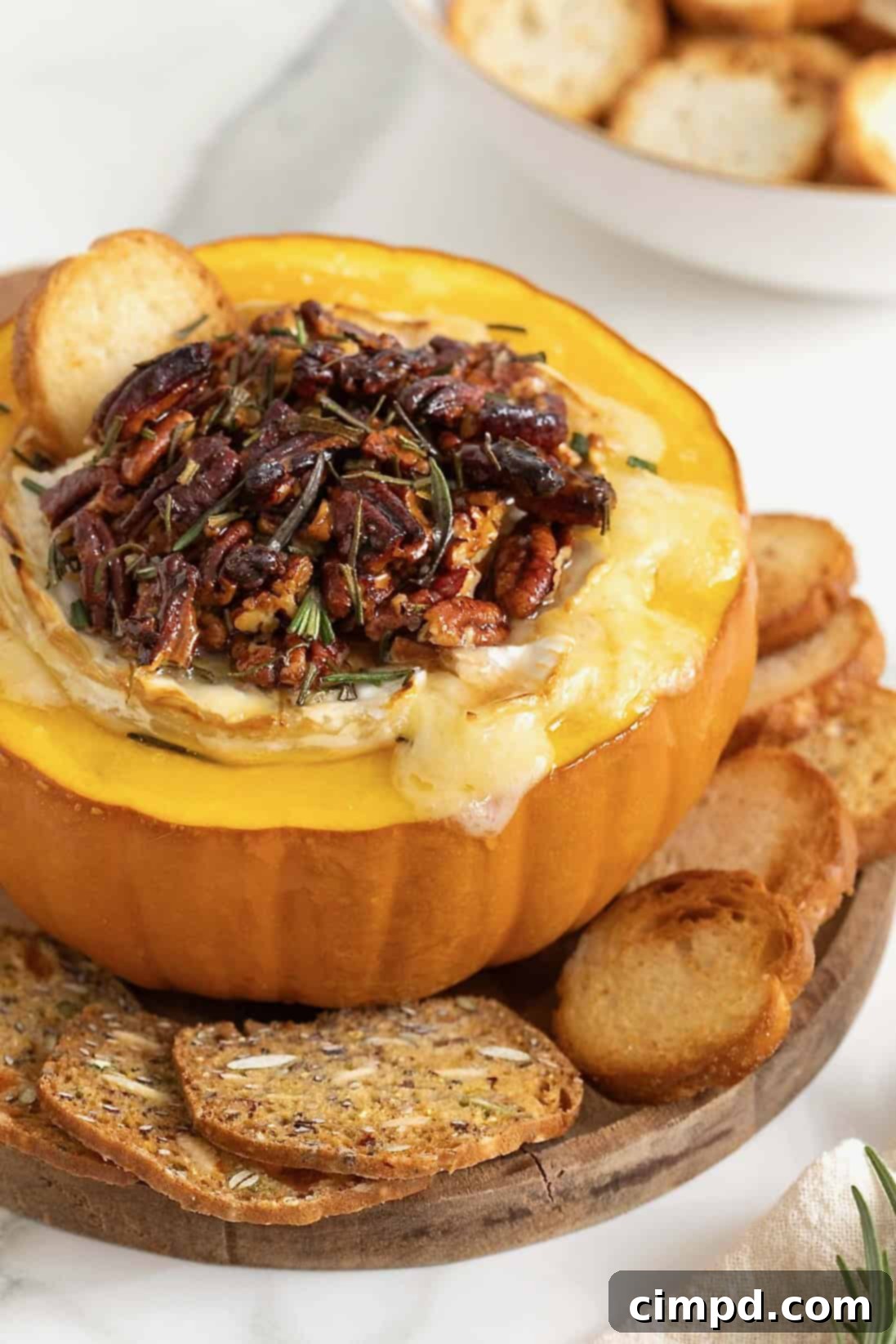 A perfectly baked brie nestled in a hollowed-out mini pumpkin, garnished with glistening maple pecans and vibrant rosemary sprigs, ready to be served.