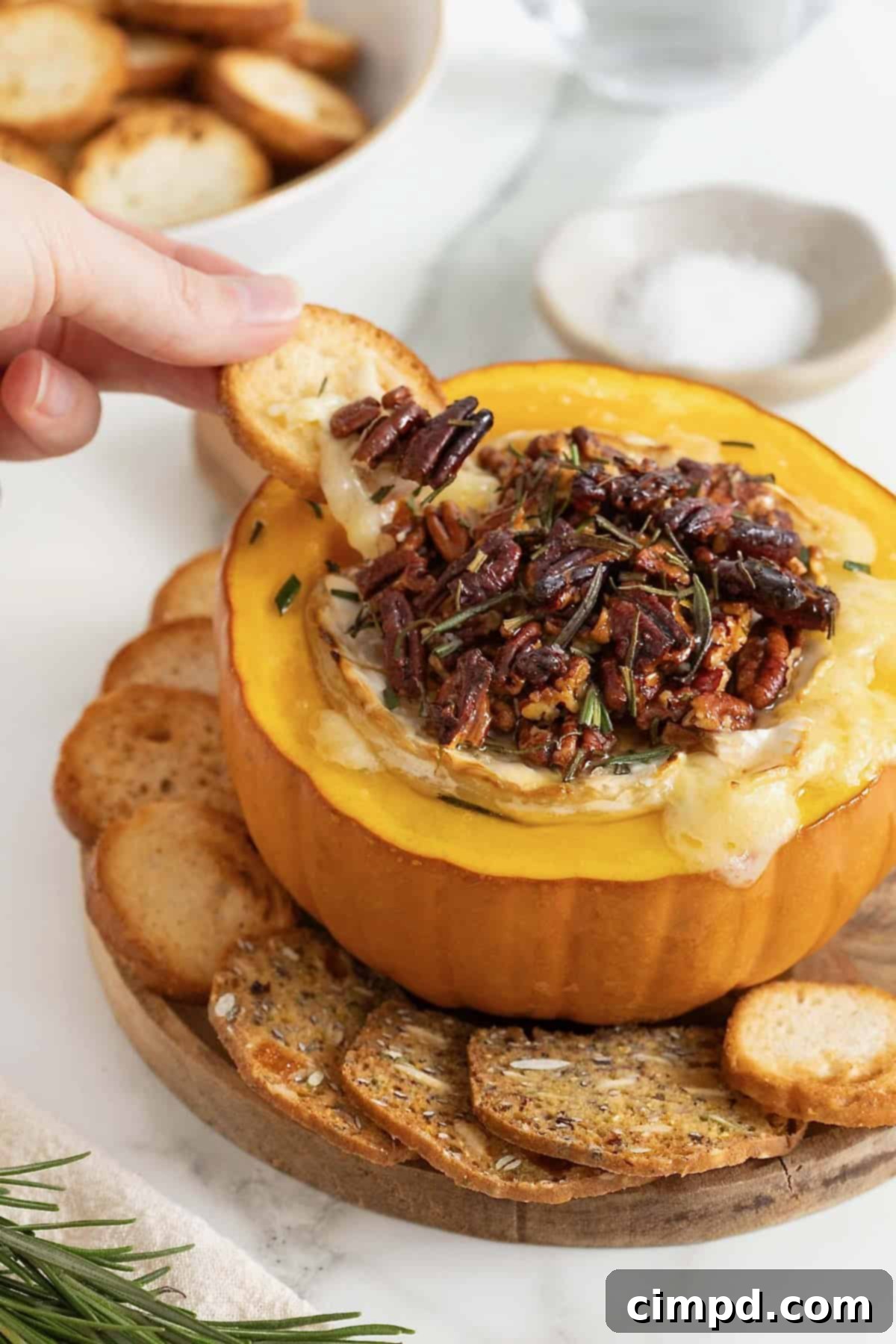 A hand dipping a cracker into the warm, gooey baked brie, served within a mini pumpkin, surrounded by an assortment of crackers and crostini on a rustic wooden board.