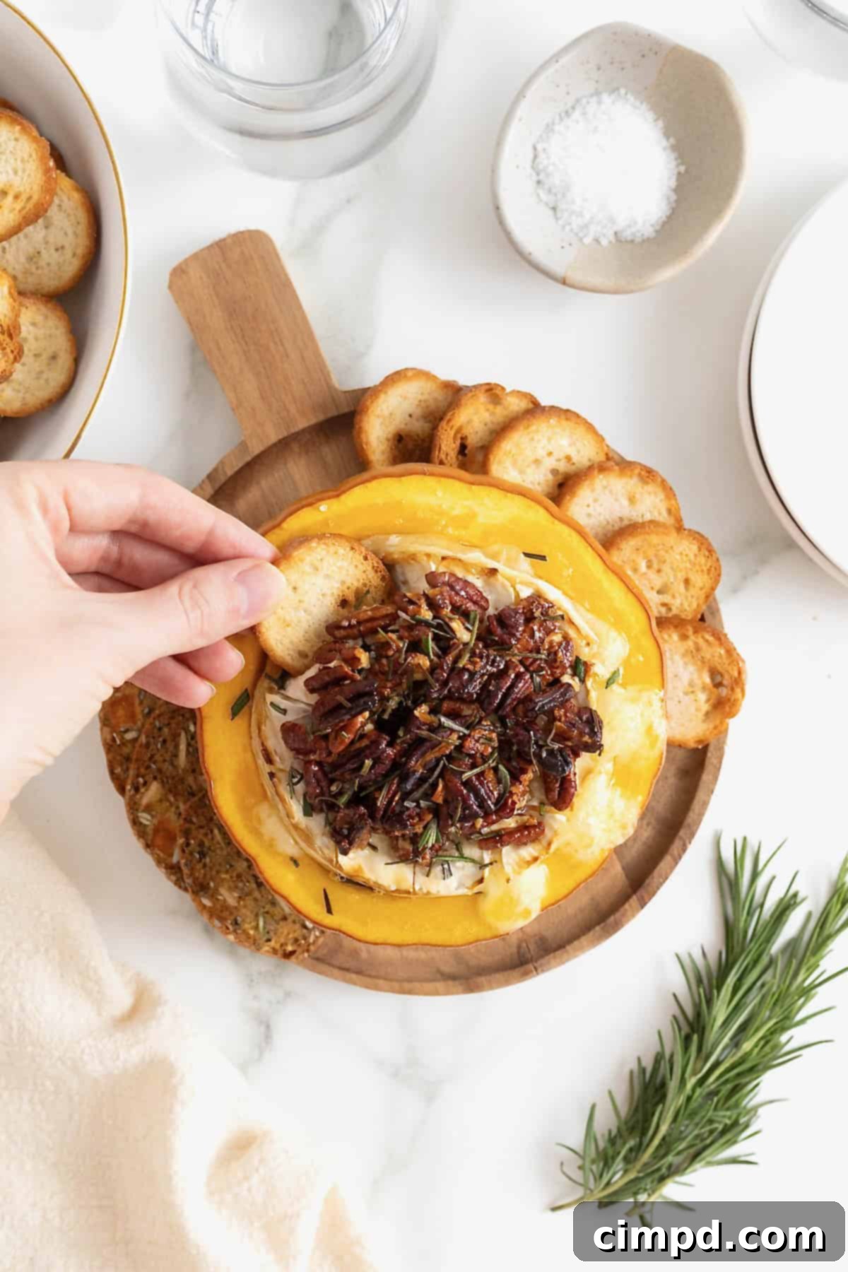 Another close-up shot of a cracker being dipped into the warm, melty baked brie, nestled in its pumpkin vessel, surrounded by various dipping options.