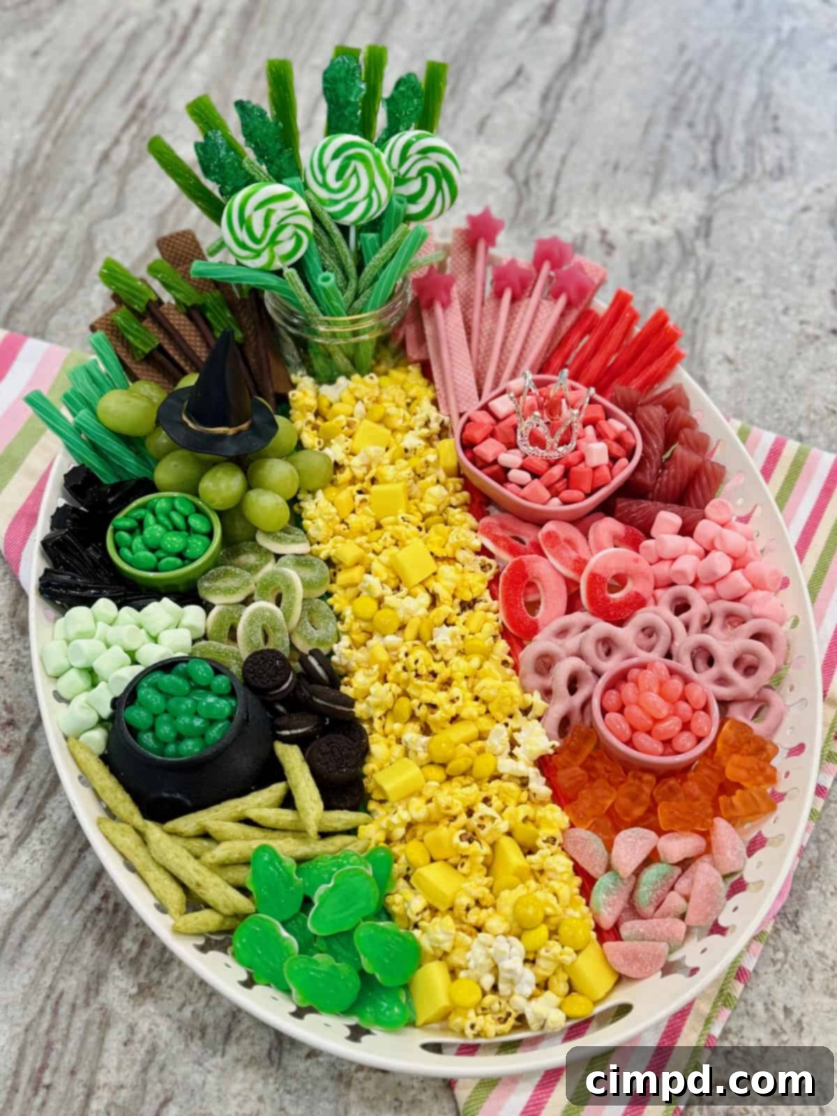 A vibrant snack board themed after the Wicked movie, featuring green and pink candies, a yellow brick road, and various treats, set on a gray marble counter.