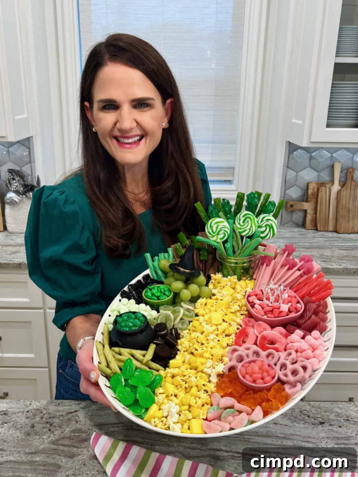 Maegan Brown, the BakerMama, smiling and holding the complete Wicked Movie Snack Board, wearing a dark green sweater.