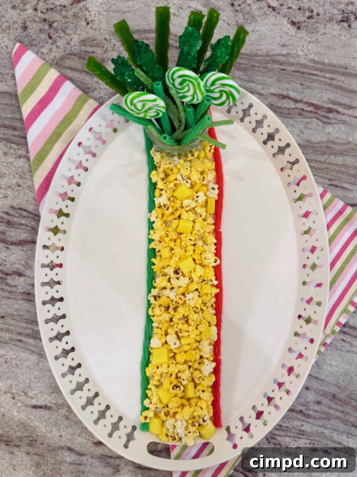 An oblong white serving tray with a prominent yellow column of buttered popcorn and yellow candies, leading to a jar of green treats at the top.
