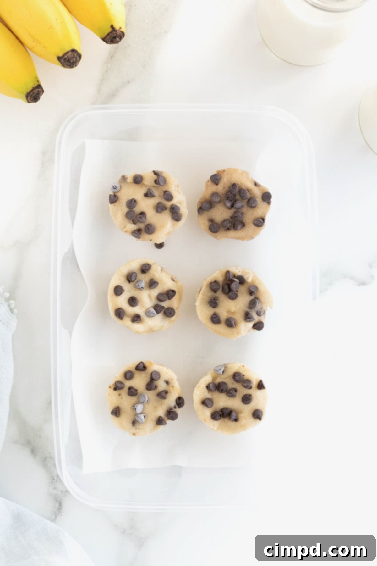 Six perfectly formed banana ice cream bites, speckled with chocolate chips, neatly arranged in a freezer-safe plastic container with parchment paper between layers.