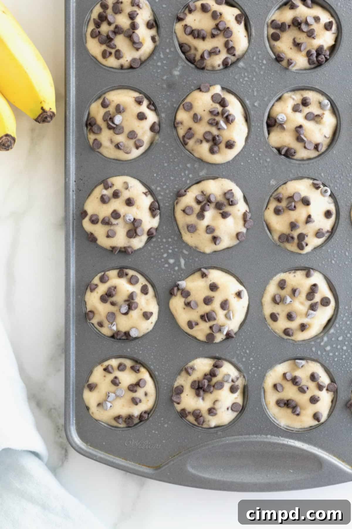 An aluminum mini muffin pan filled with Two-Ingredient Banana Chocolate Chip Ice Cream Bites, garnished with extra chips, resting on a pristine white marble counter.