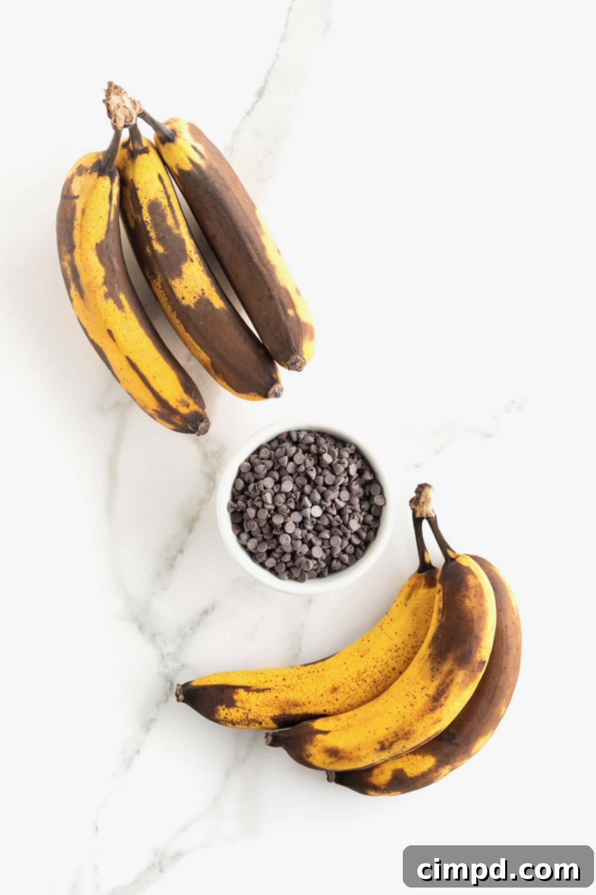 Two generous bunches of perfectly overripe bananas flank a small white dish brimming with mini chocolate chips, set against a rustic wooden backdrop.