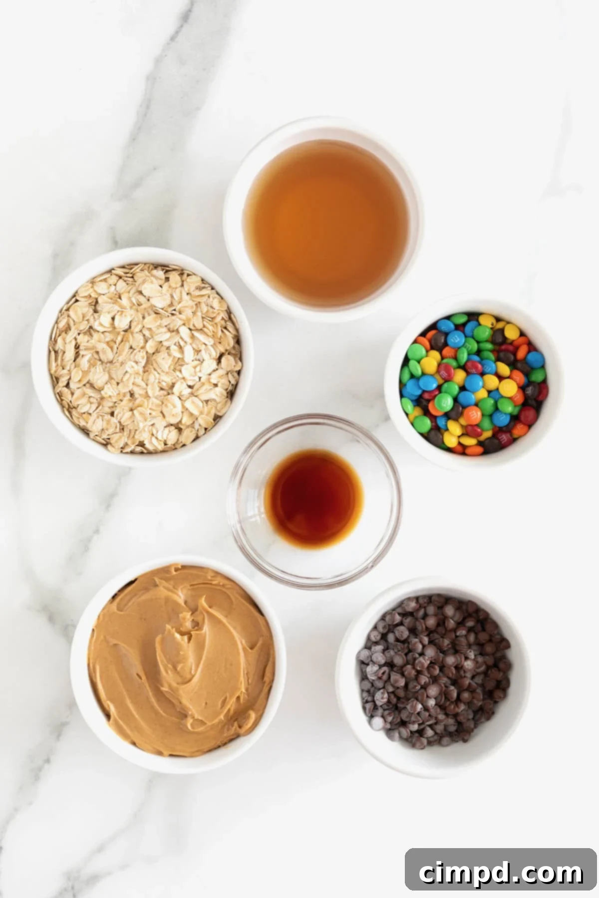 No-Bake Monster Cookie Bites 7 A vibrant arrangement of all the ingredients needed for no-bake monster cookie balls, neatly displayed in small white and glass dishes on a white marble counter.