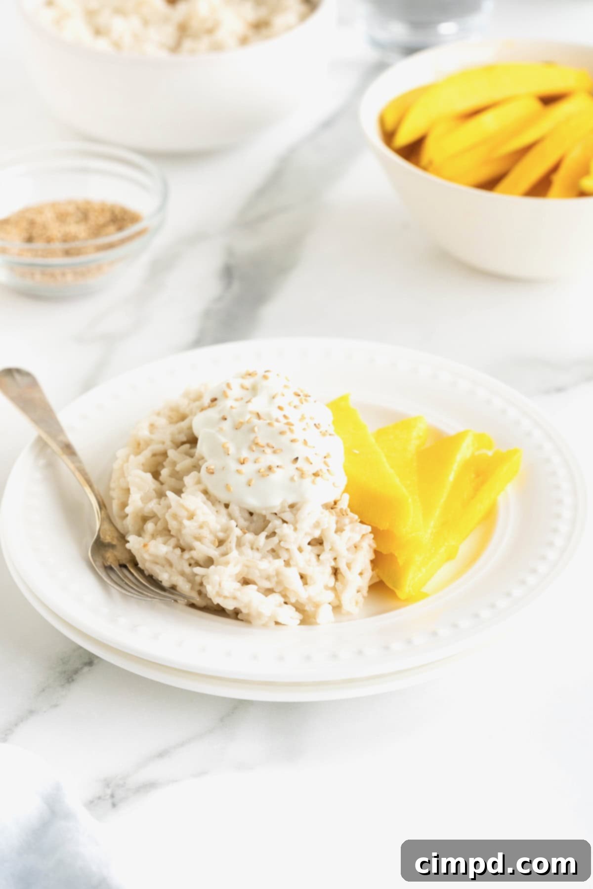A small white plate of mango sticky rice sits on a pristine white marble counter, accompanied by a tiny bowl of sesame seeds.