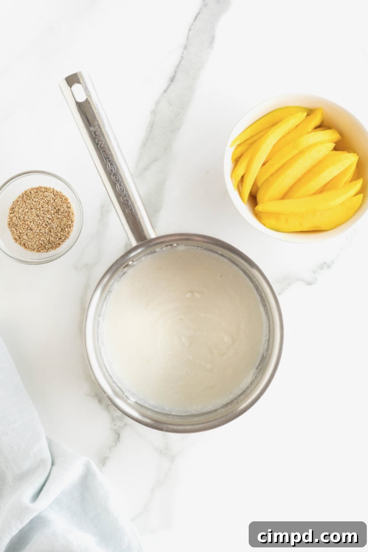 A small saucepan holds the thickening cream sauce for Thai mango sticky rice. A dish of bright sliced mango sits to the right, and a tiny dish of sesame seeds to the left.
