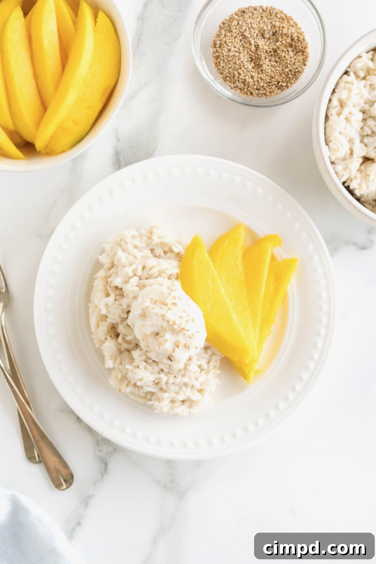 A small white plate displays slices of fresh mango alongside sticky rice, topped with creamy sauce and a sprinkle of toasted sesame seeds.