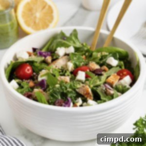 A green salad with tomato, feta, and quinoa in a large white bowl with two gold tone spoon handles sticking out.