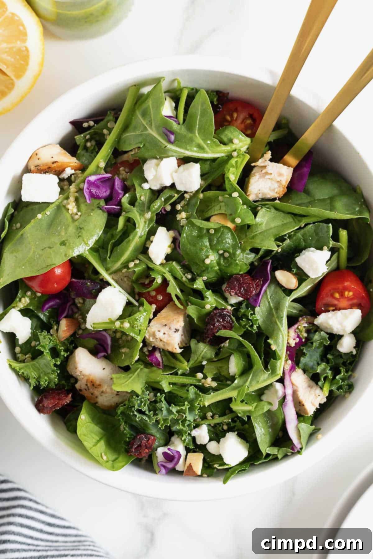 A generous portion of green salad, complete with vibrant cherry tomatoes, fluffy quinoa, and crumbled feta cheese, presented in a large white bowl with two golden spoon handles.