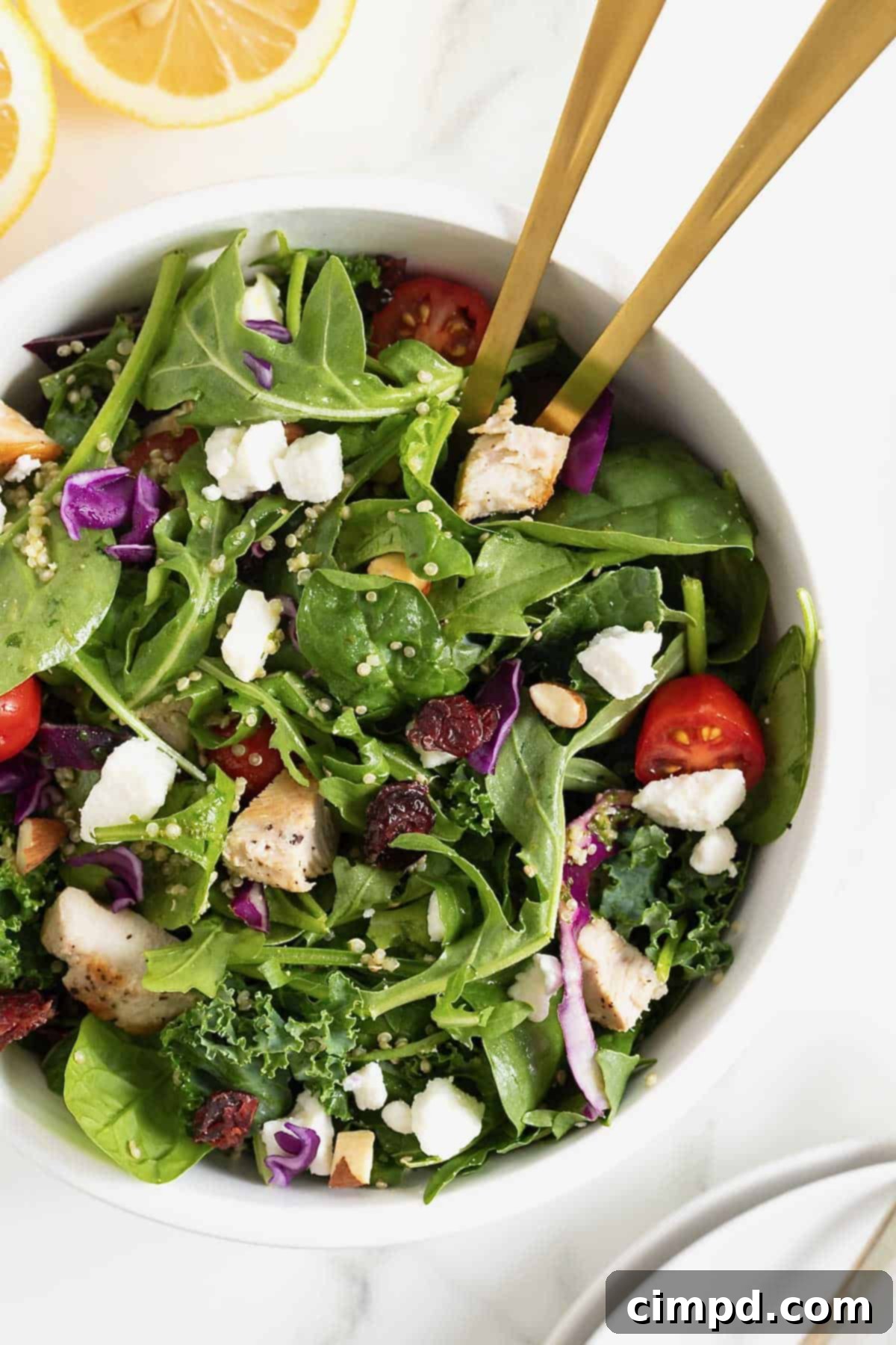 A beautifully arranged green salad, featuring cherry tomatoes, quinoa, and crumbled feta, served in a large white bowl with two golden spoon handles for serving.