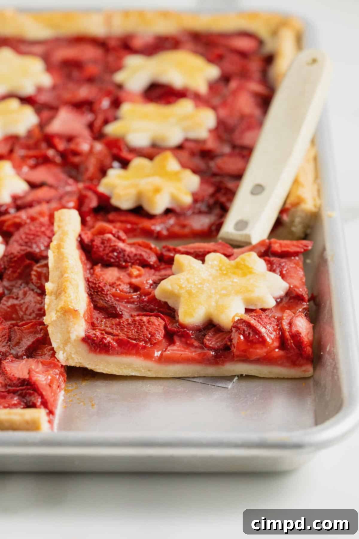 Summer Strawberry Slab Pie 4 A delicious Strawberry Slab Pie, fresh from the oven, baked in an aluminum rimmed baking sheet. A serving spatula lifts a piece, showcasing the golden-brown crust and bubbling strawberry filling.