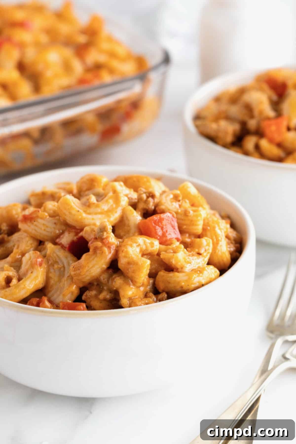 Fiesta Mac and Cheese 5 Two white bowls of Mexi Mac and Cheese, with a fork resting beside the front bowl. A 9x13 glass casserole dish is blurred in the background.