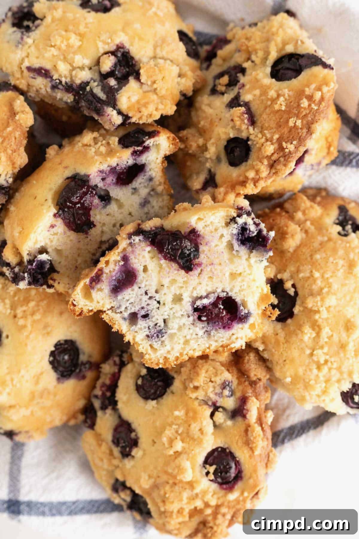 A blueberry muffin cut in half, revealing the soft, airy interior speckled with blueberries, next to a pile of whole muffins.