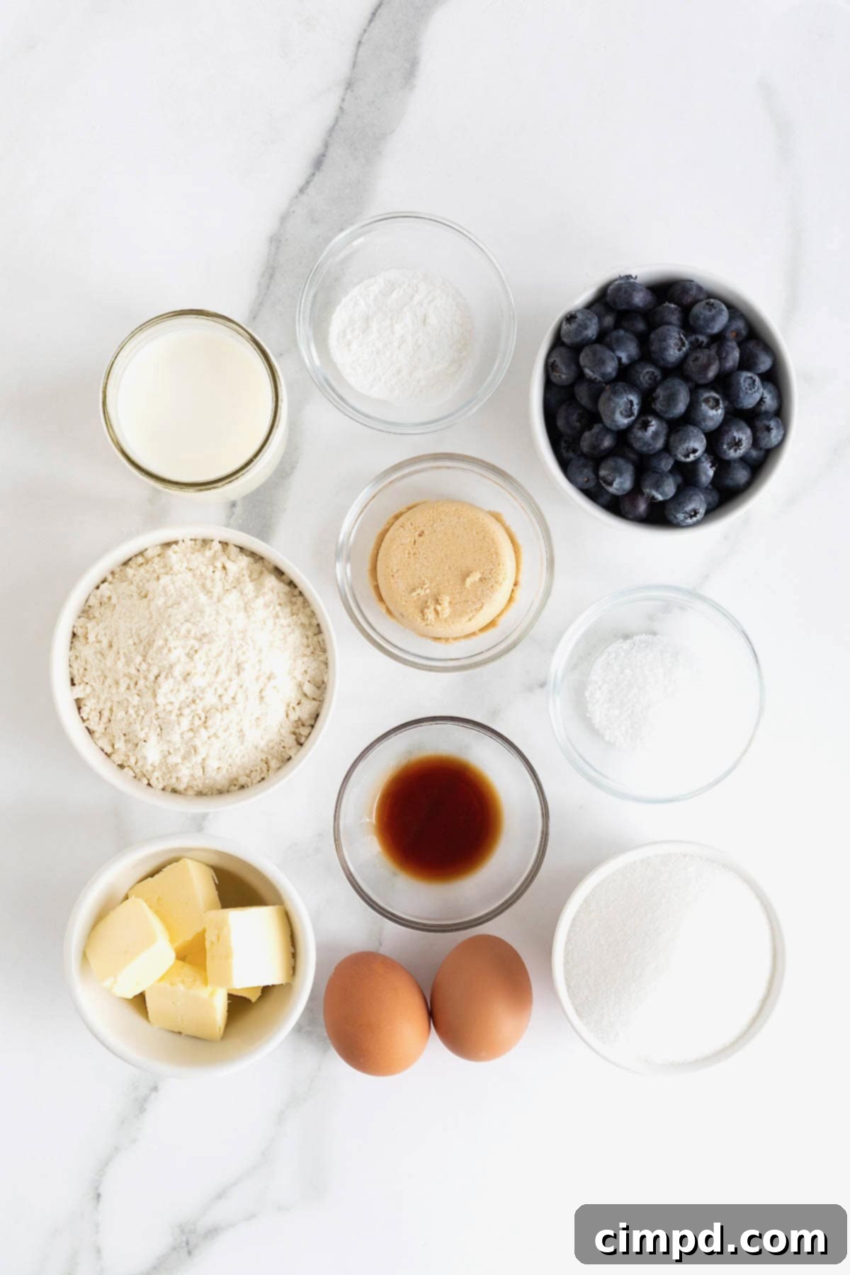 Various ingredients for blueberry streusel muffins laid out in small glass dishes on a white marble counter, ready for preparation.