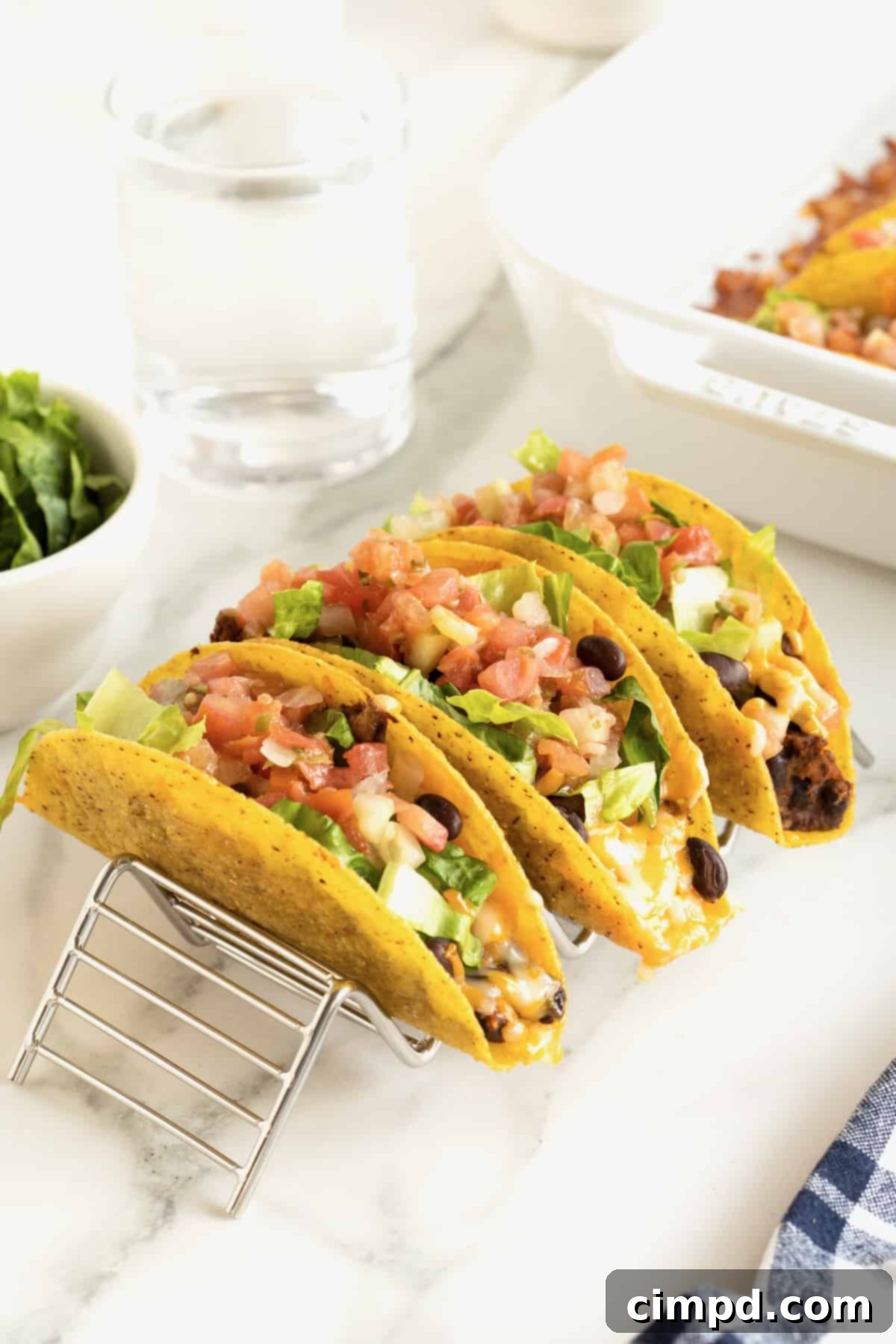 Three perfectly baked turkey and black bean tacos, topped with fresh diced tomatoes and crisp lettuce, elegantly displayed in an acrylic taco stand on a dining table.