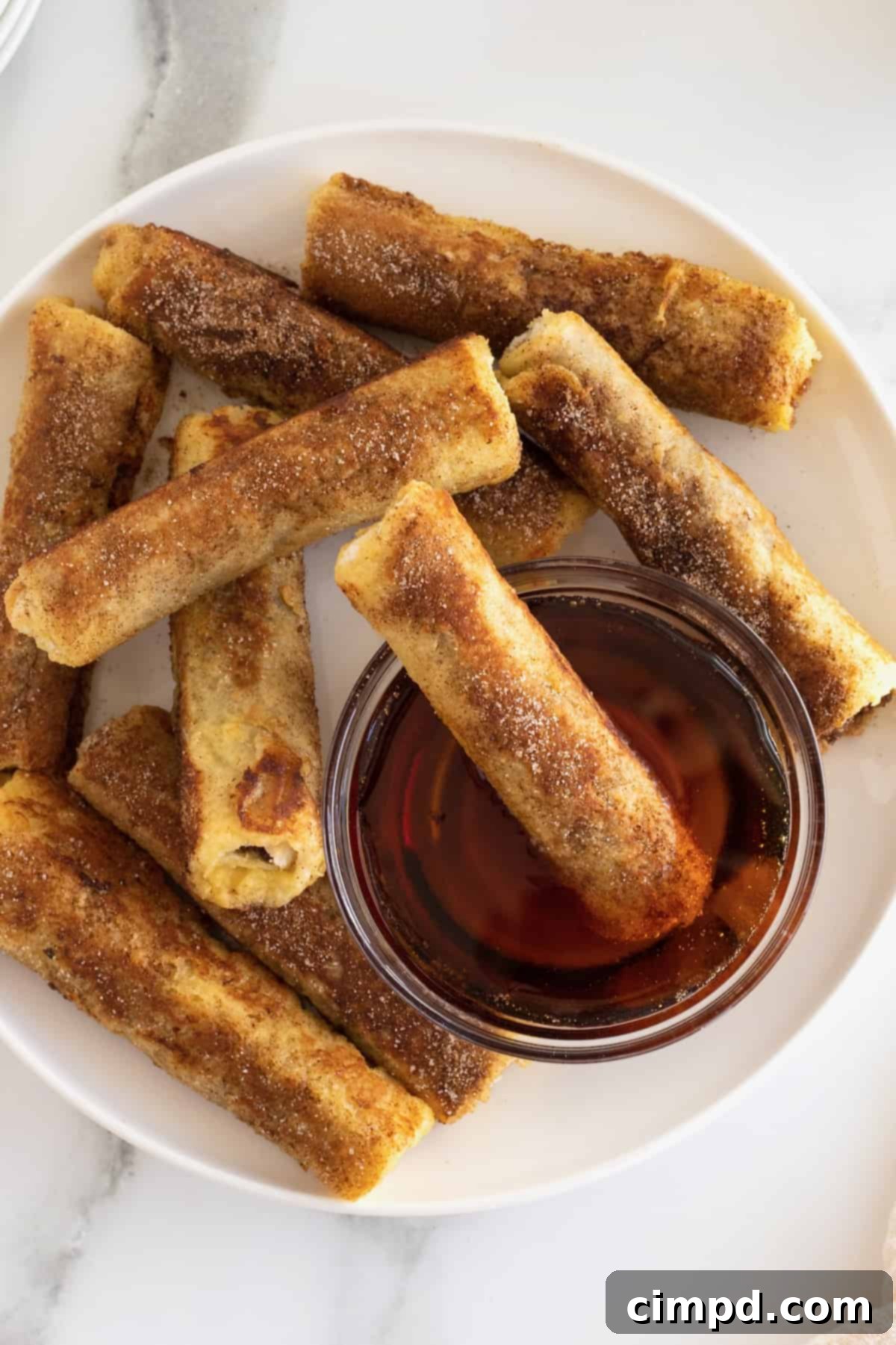 Golden brown cinnamon sugar French toast roll-ups elegantly arranged around a glass bowl of warm, amber maple syrup.