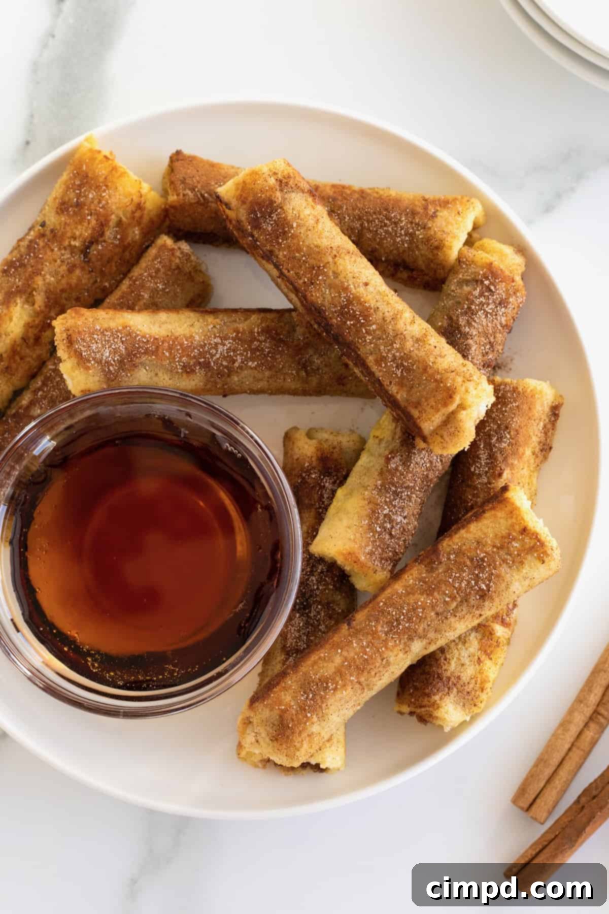 A vibrant display of cinnamon sugar-covered French toast roll-ups elegantly arranged around a glass bowl overflowing with warm, amber maple syrup.