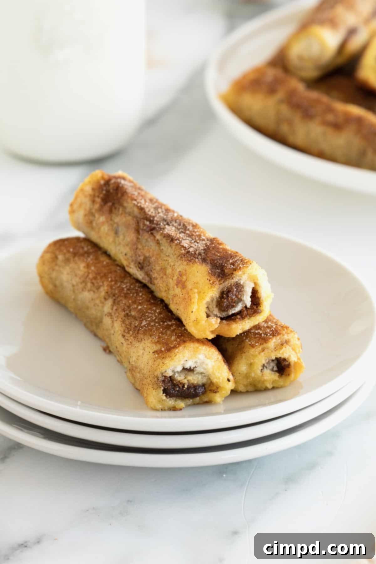 A neat pile of three French toast roll-ups rests on a stack of three pristine white plates, highlighting their delicious appearance.