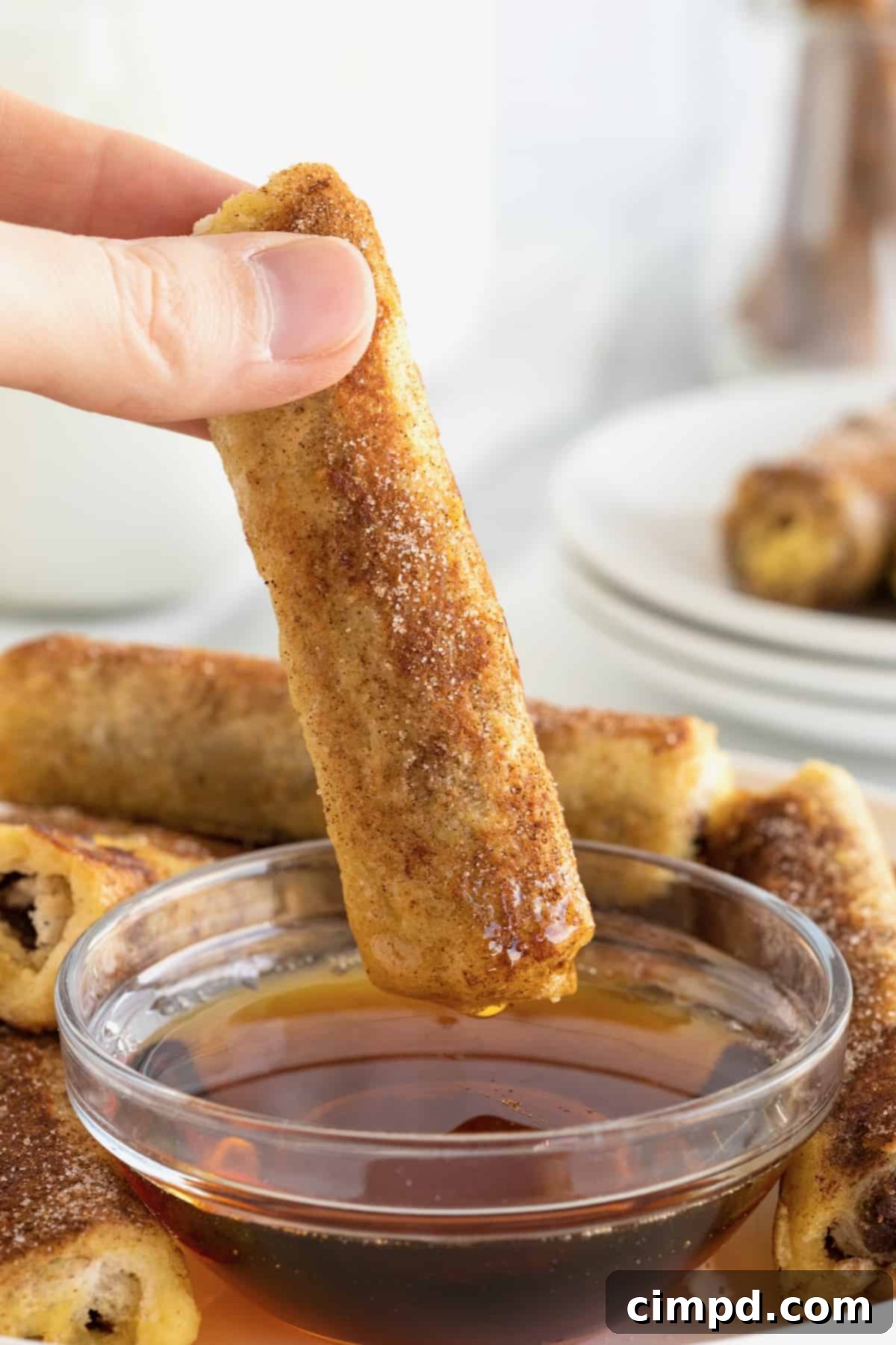A hand gently holds a perfectly rolled French toast roll-up, poised above a glistening glass bowl of rich maple syrup, ready for a delectable dip.