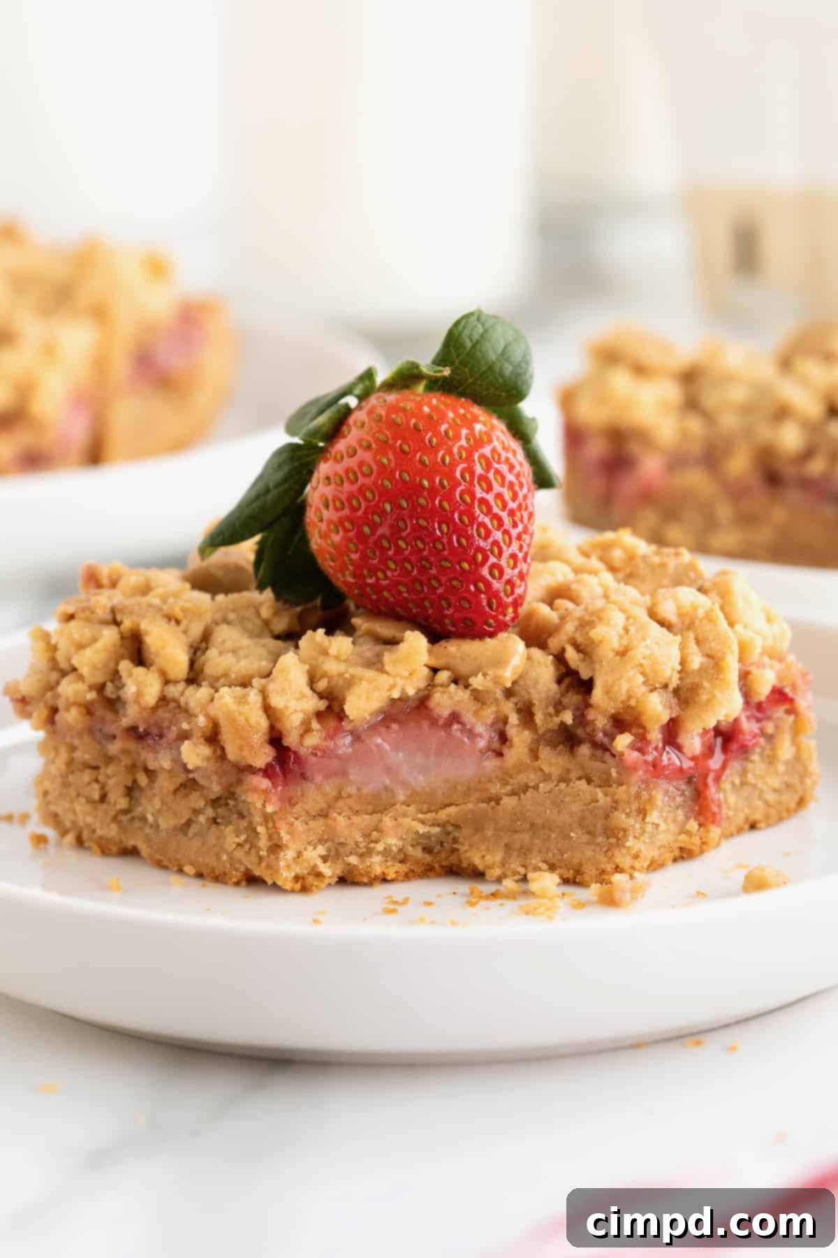Strawberry Peanut Butter Swirl Bars 3 A strawberry peanut butter bar on a white rimmed plate with a bite taken out, showcasing the baked-in strawberry pieces. A whole fresh strawberry sits delicately on top.