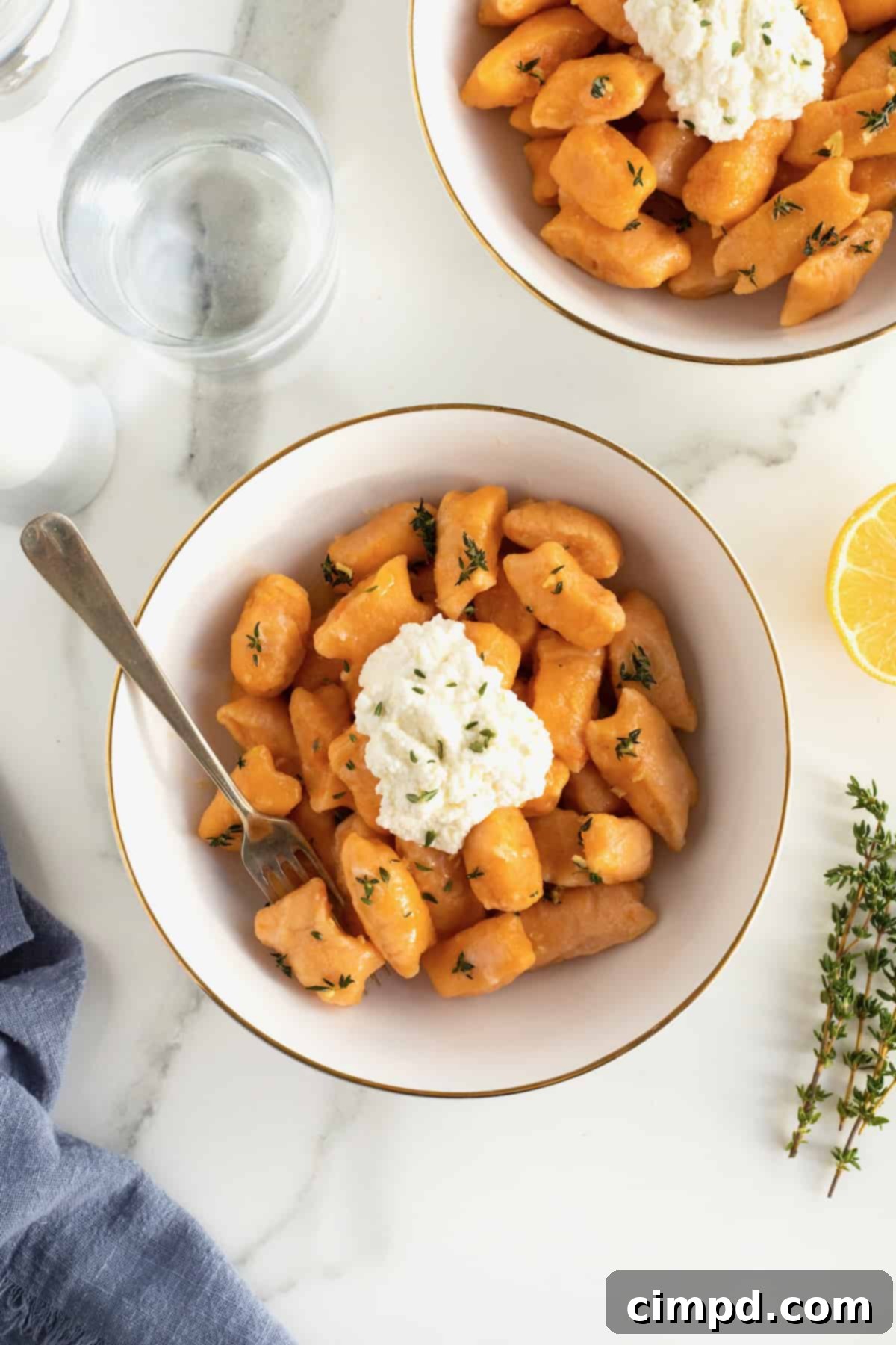 A gold-rimmed white bowl brimming with perfectly cooked sweet potato gnocchi, topped with a creamy dollop of fresh ricotta and flecks of herbs.