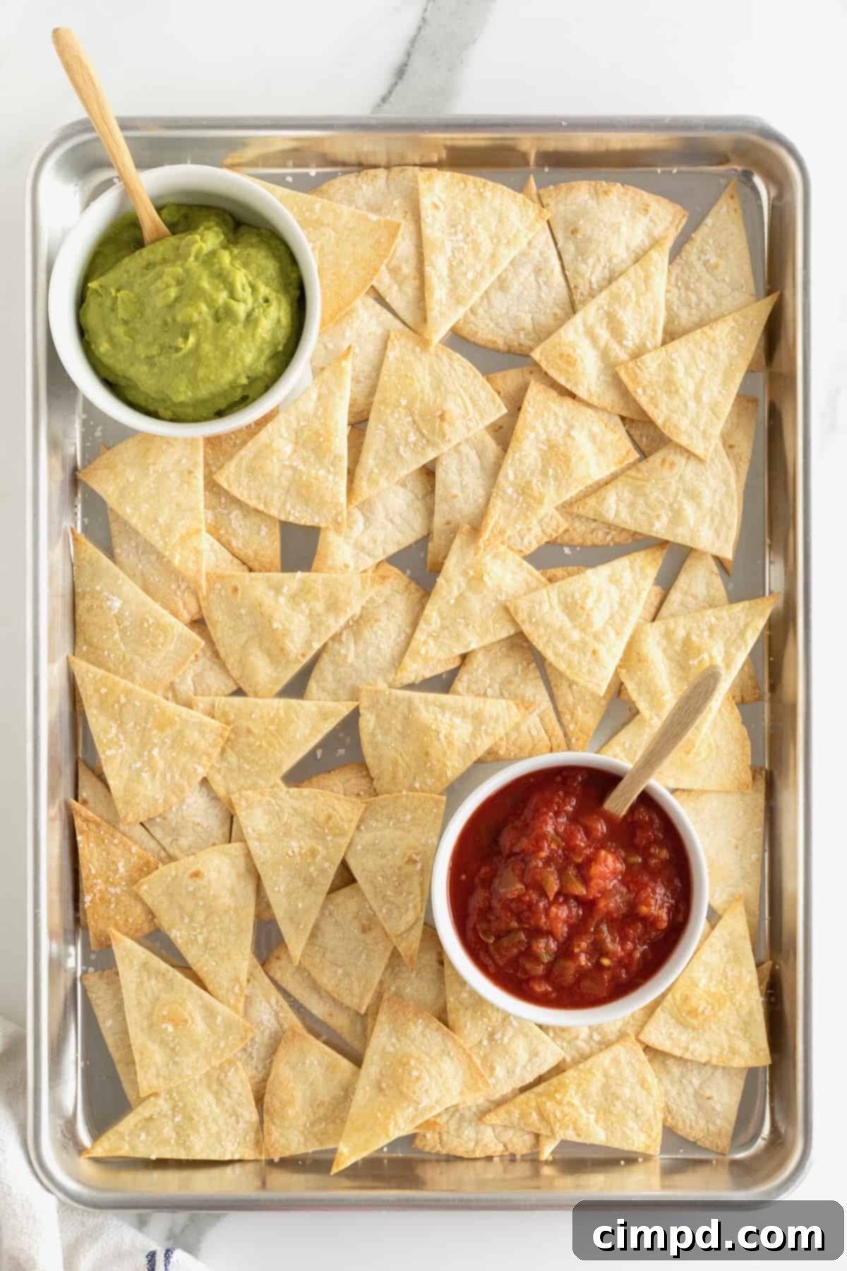 Crispy triangle-cut flour tortilla chips on a baking sheet, ready for serving, with small bowls of vibrant salsa and creamy guacamole.