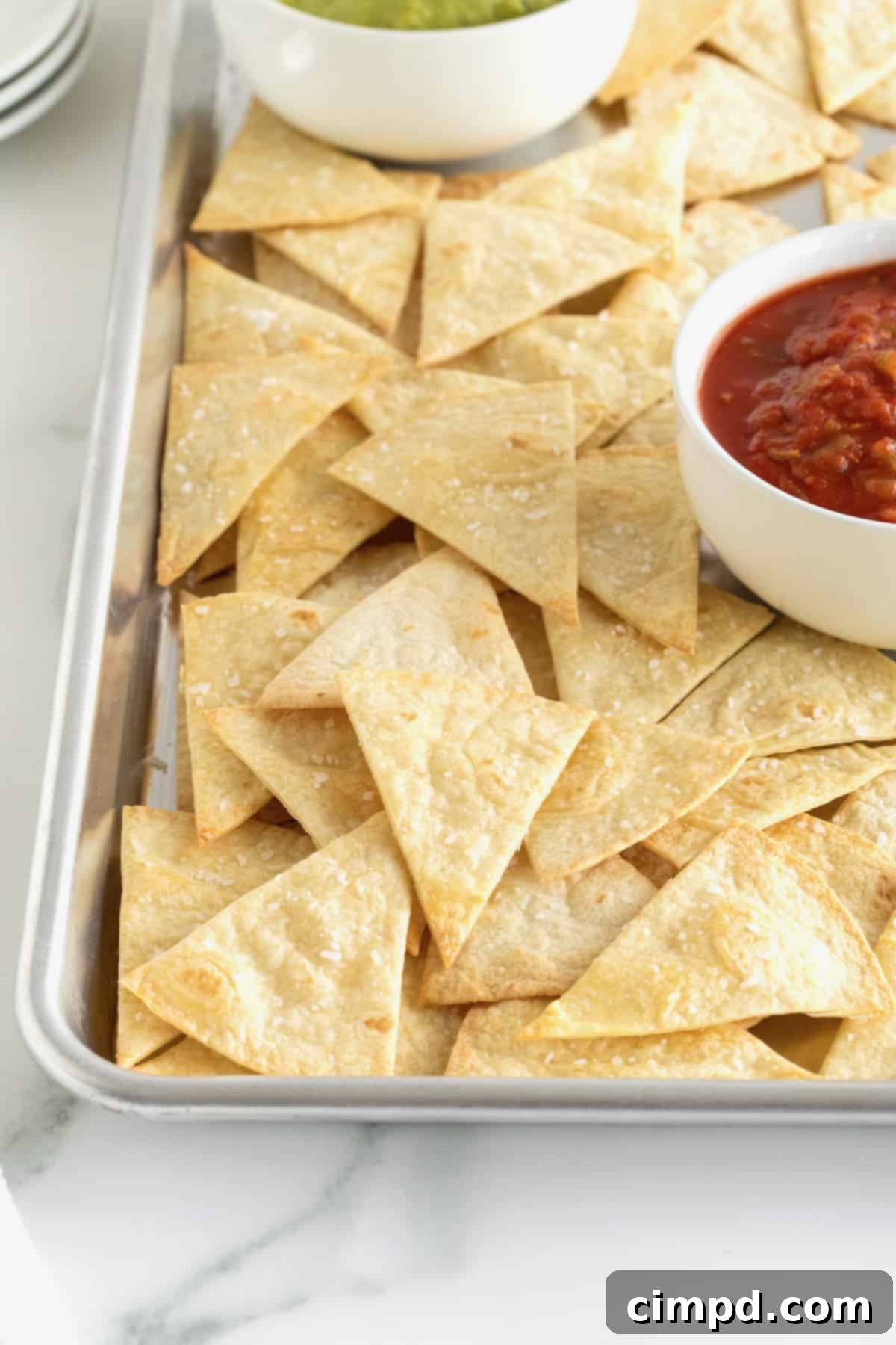 Freshly baked, golden-brown triangle-cut flour tortilla chips spread evenly on a baking sheet, with a small bowl of chunky salsa in the background.